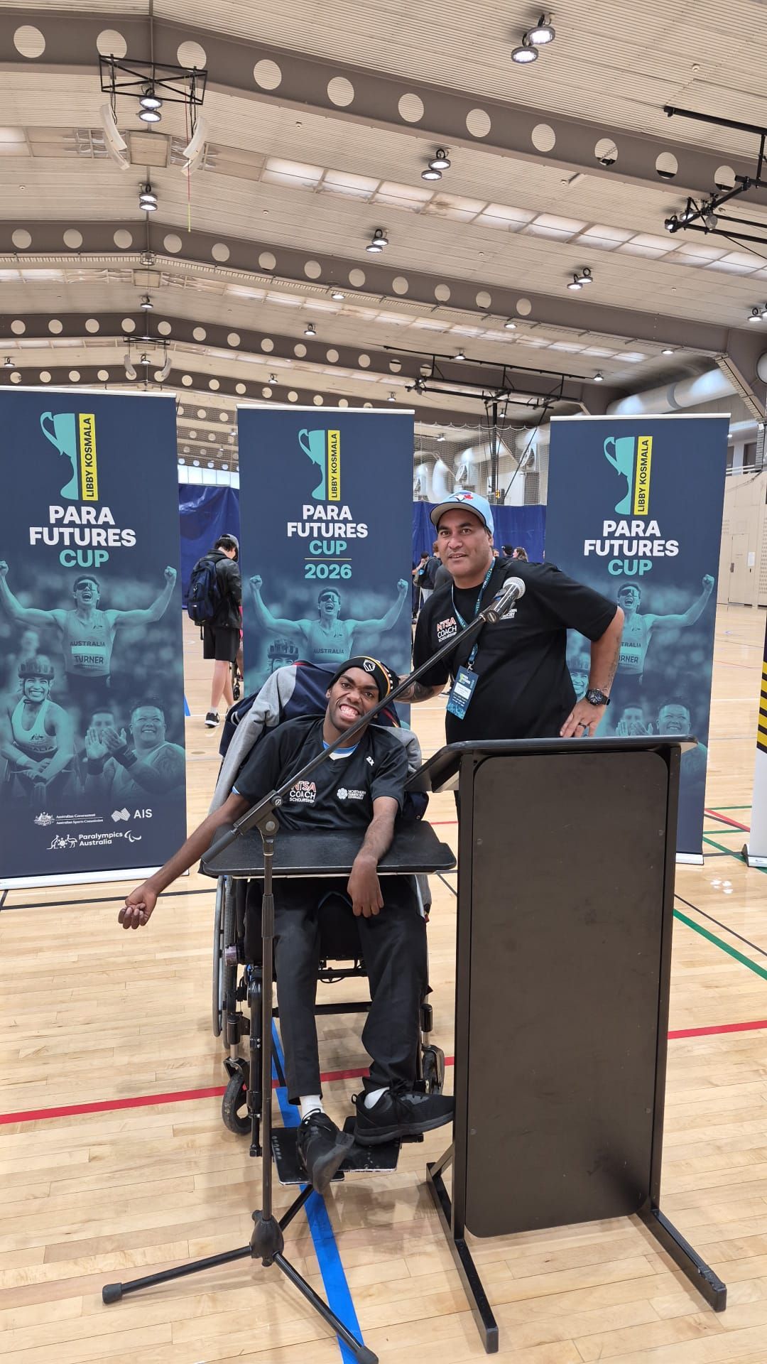 Athletes at an indoor event podium, one seated in a wheelchair, with sports banners behind them