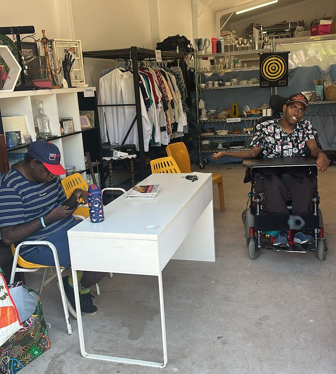 Two people in a clothing shop, one seated at a table and one in a wheelchair near clothing racks.