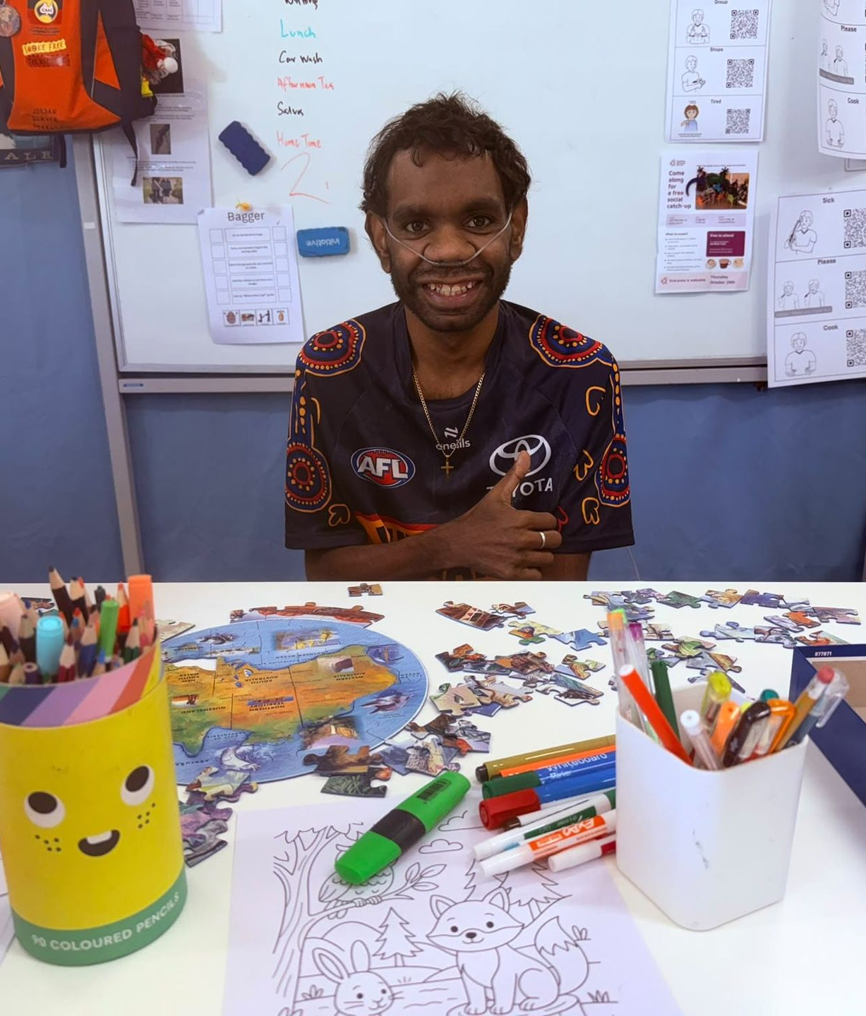 Person smiling at a desk with colored pencils, paper crafts, and a green cup in a classroom.