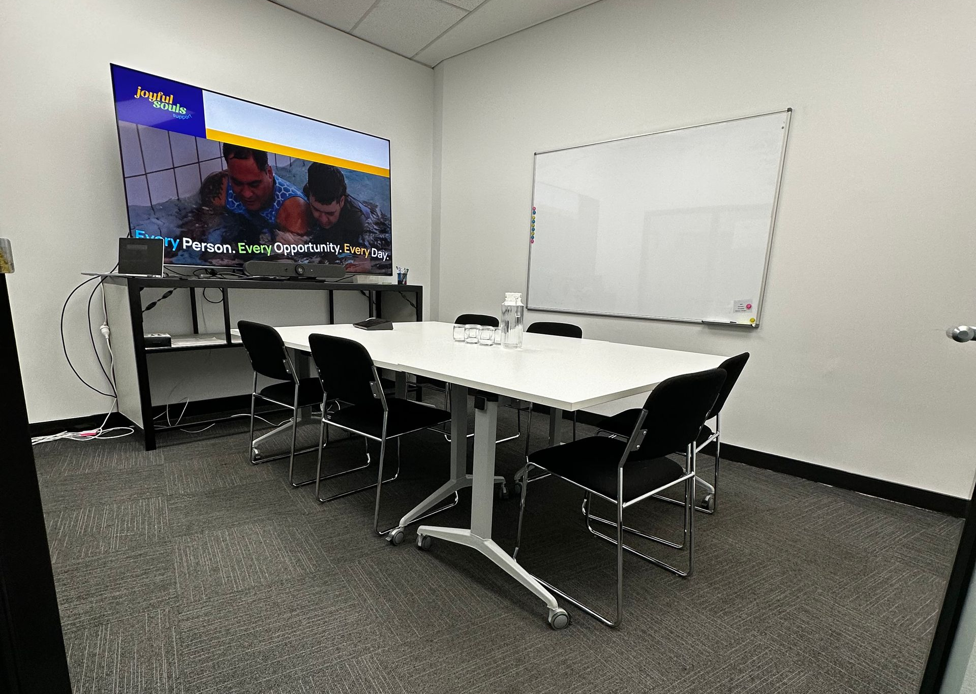 Conference room with white table, black chairs, wall-mounted TV, and whiteboard on gray carpet