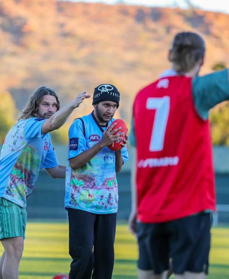 Three people in sports jerseys on a field, two clapping toward a player in red