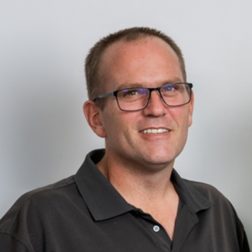 Man wearing glasses and a dark polo shirt, smiling against a plain light background