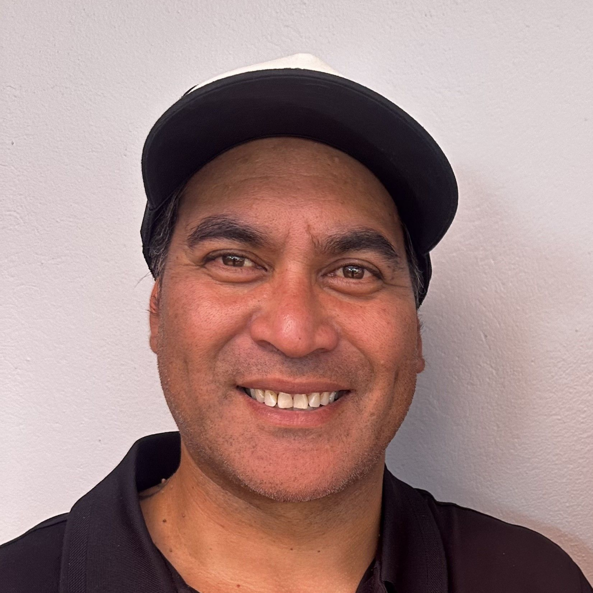 Shannon miling person in a black cap and dark polo shirt against a light wall.