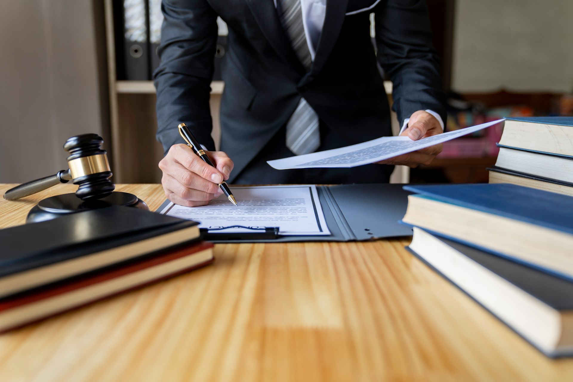 A professional in a suit signs a legal document at a desk with a gavel and stacks of books.