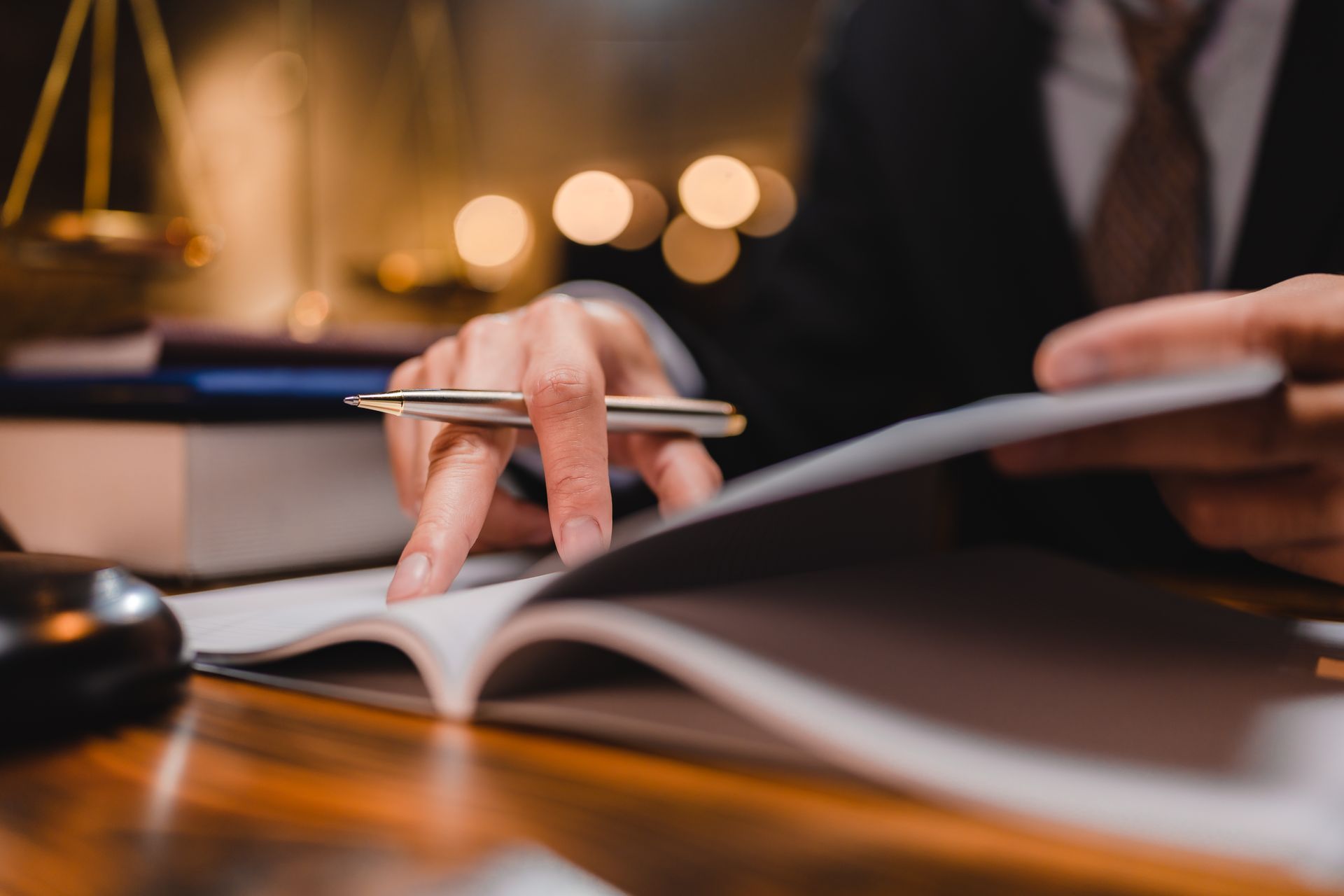 A person in a suit sits at a desk with a book, holding a pen over the open pages, with a scale of justice in the background.