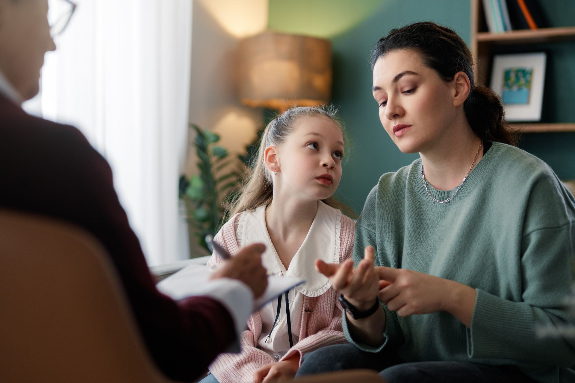 A professional sits with a child and parent in an office, documenting their conversation as the parent gestures to speak.