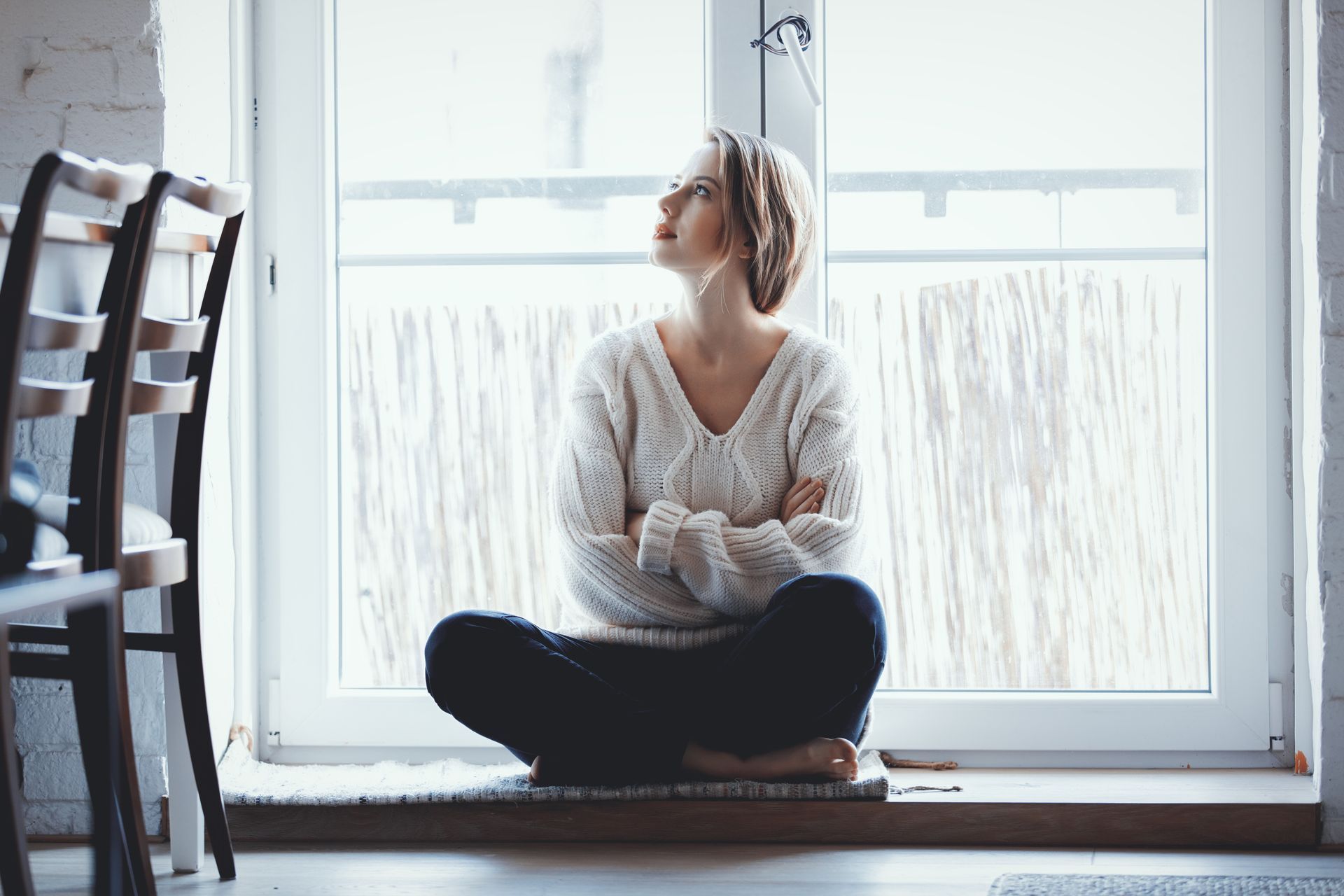 A person in a white knit sweater sitting cross-legged on a windowsill, looking upward with arms crossed.