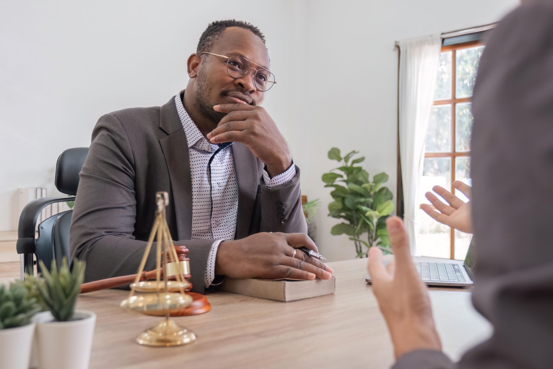 A lawyer sits at a desk with a scale of justice, listening to a client speak in a professional office.