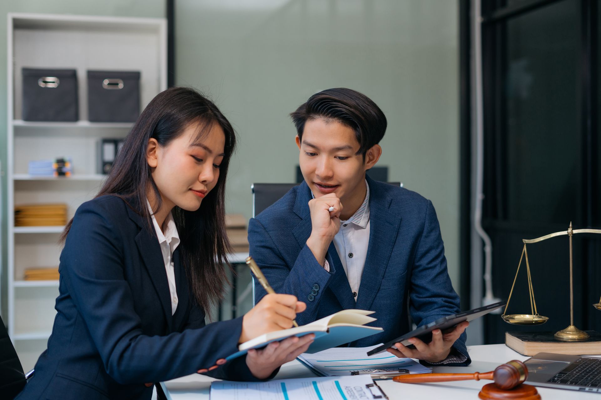 Two colleagues in business suits confer in an office, reviewing documents and a tablet with a scale of justice nearby.