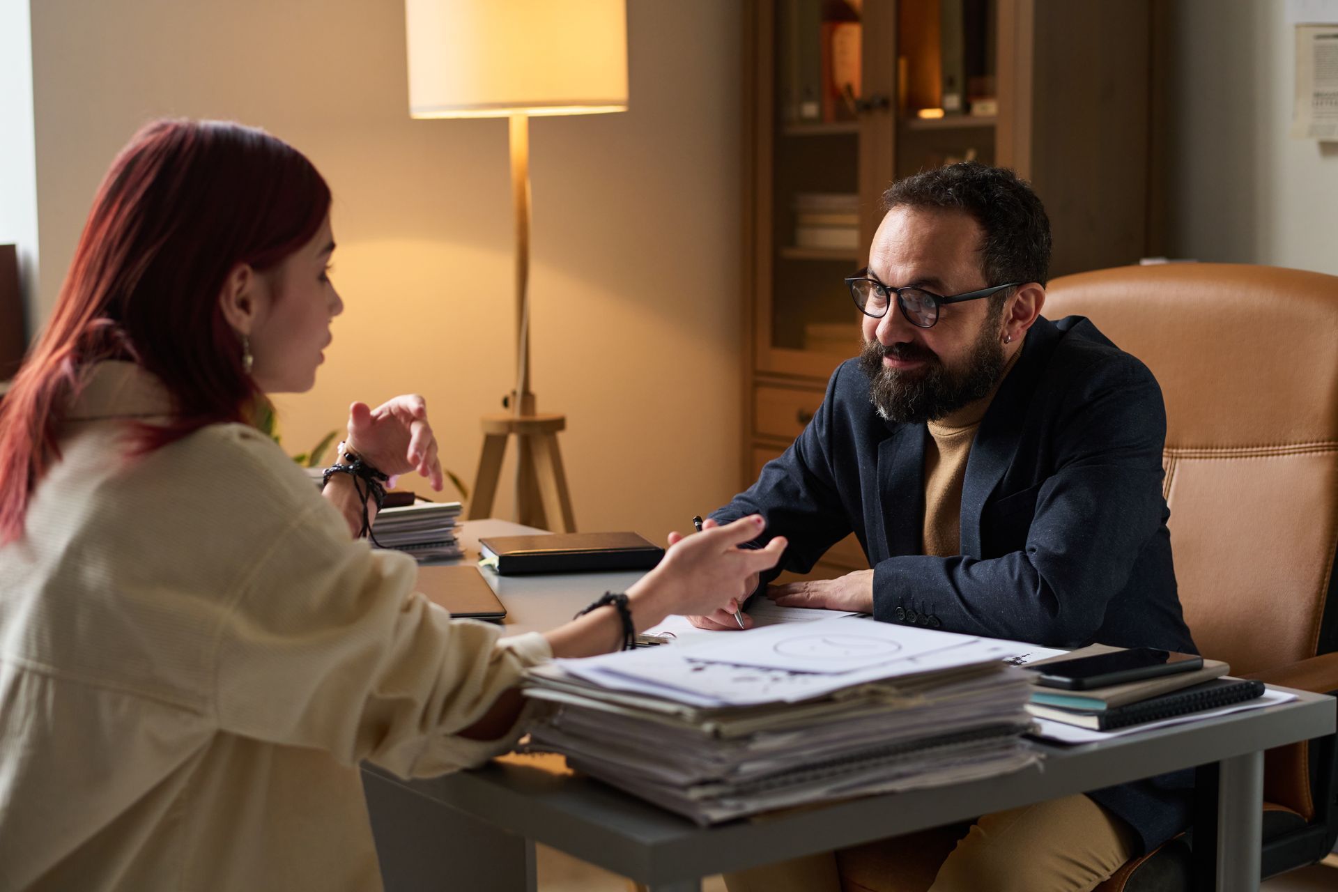 A person with red hair gestures while talking to a bearded man in glasses across a desk piled with papers in an office.