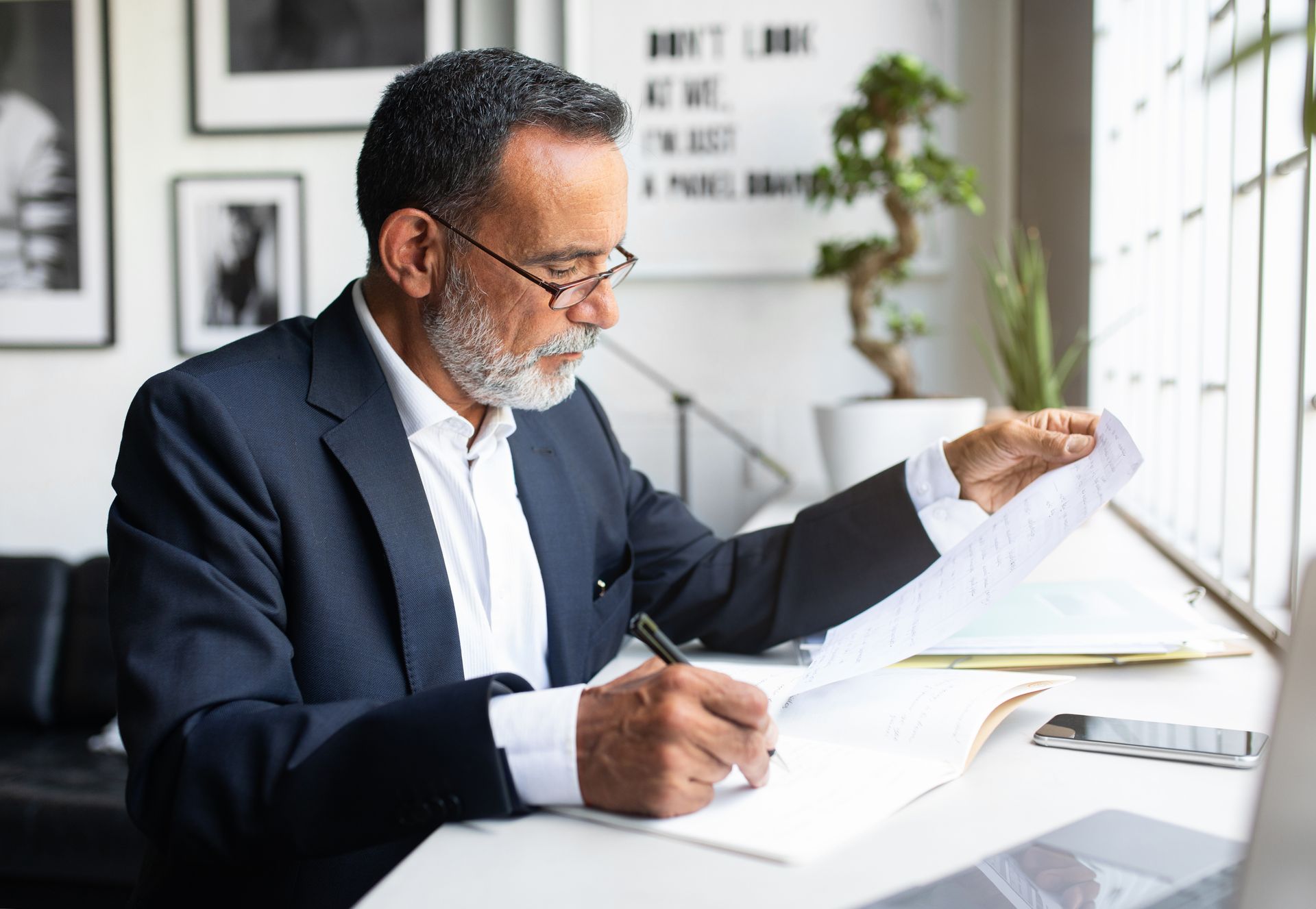 A person in a suit reviewing a document and writing in a notebook at a sunlit desk.