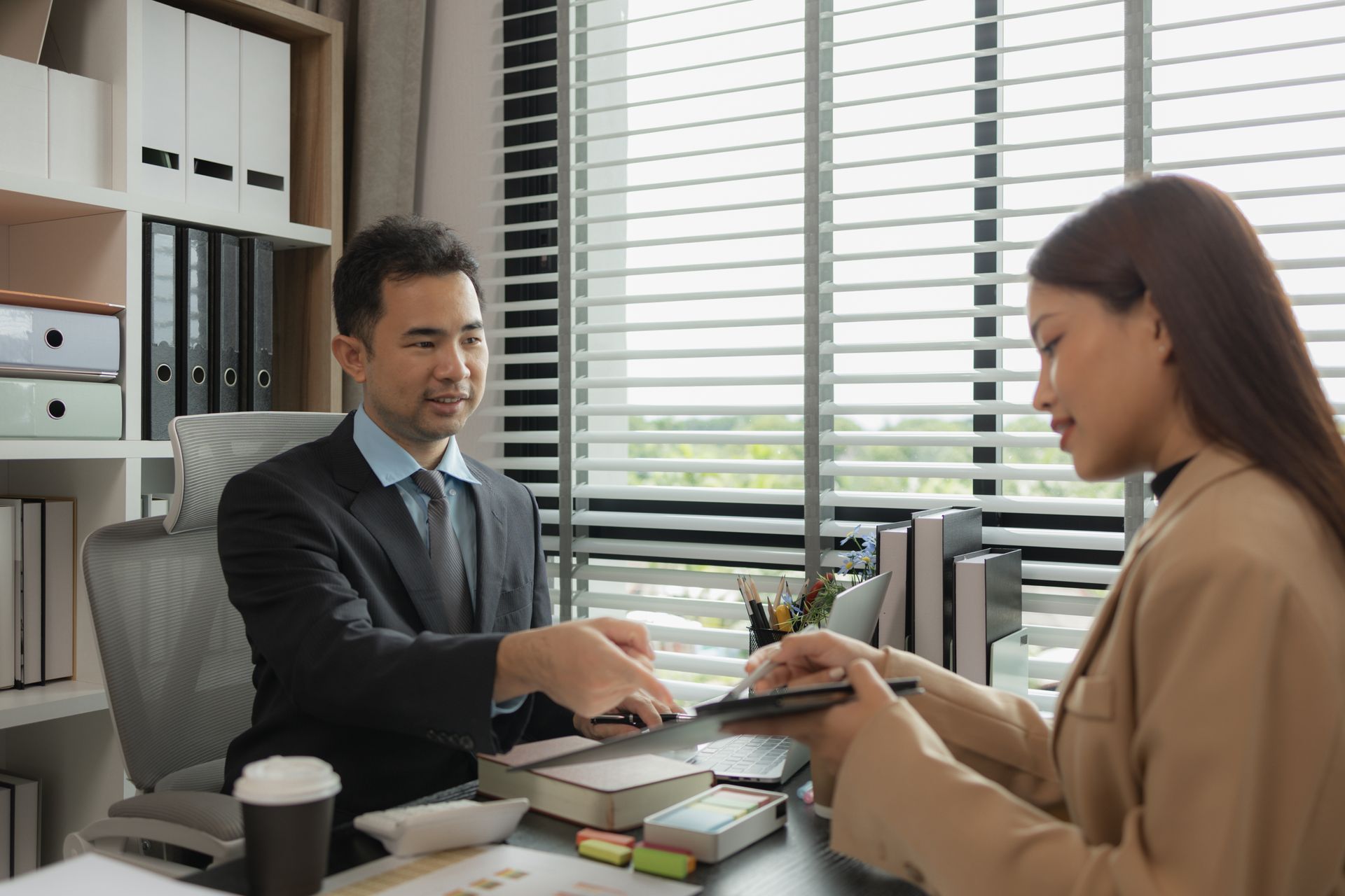 A professional sitting at a desk discusses documents with a colleague in an office with window blinds and bookshelves.