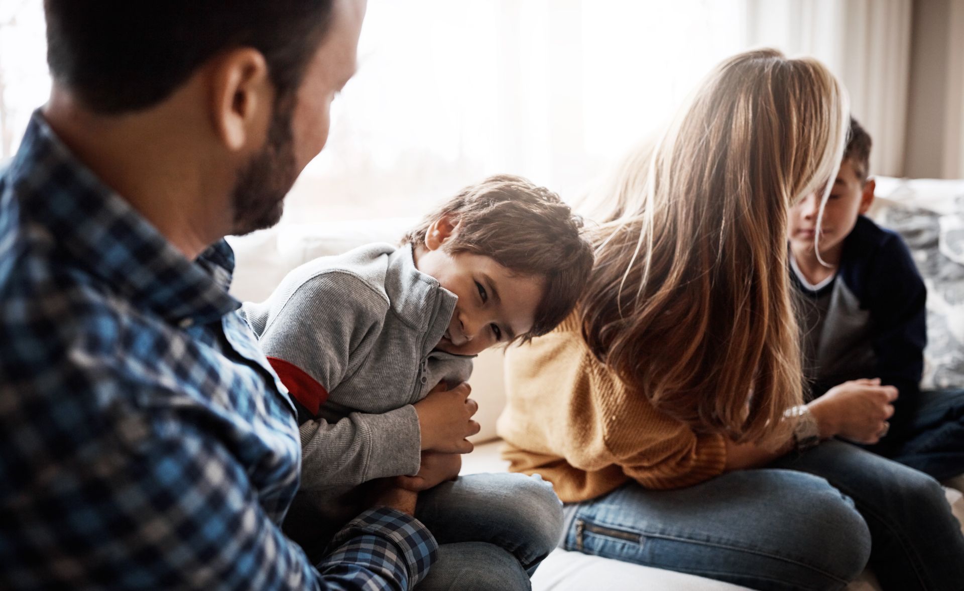 A family sits closely together indoors, with a parent comforting a child while another child looks on.