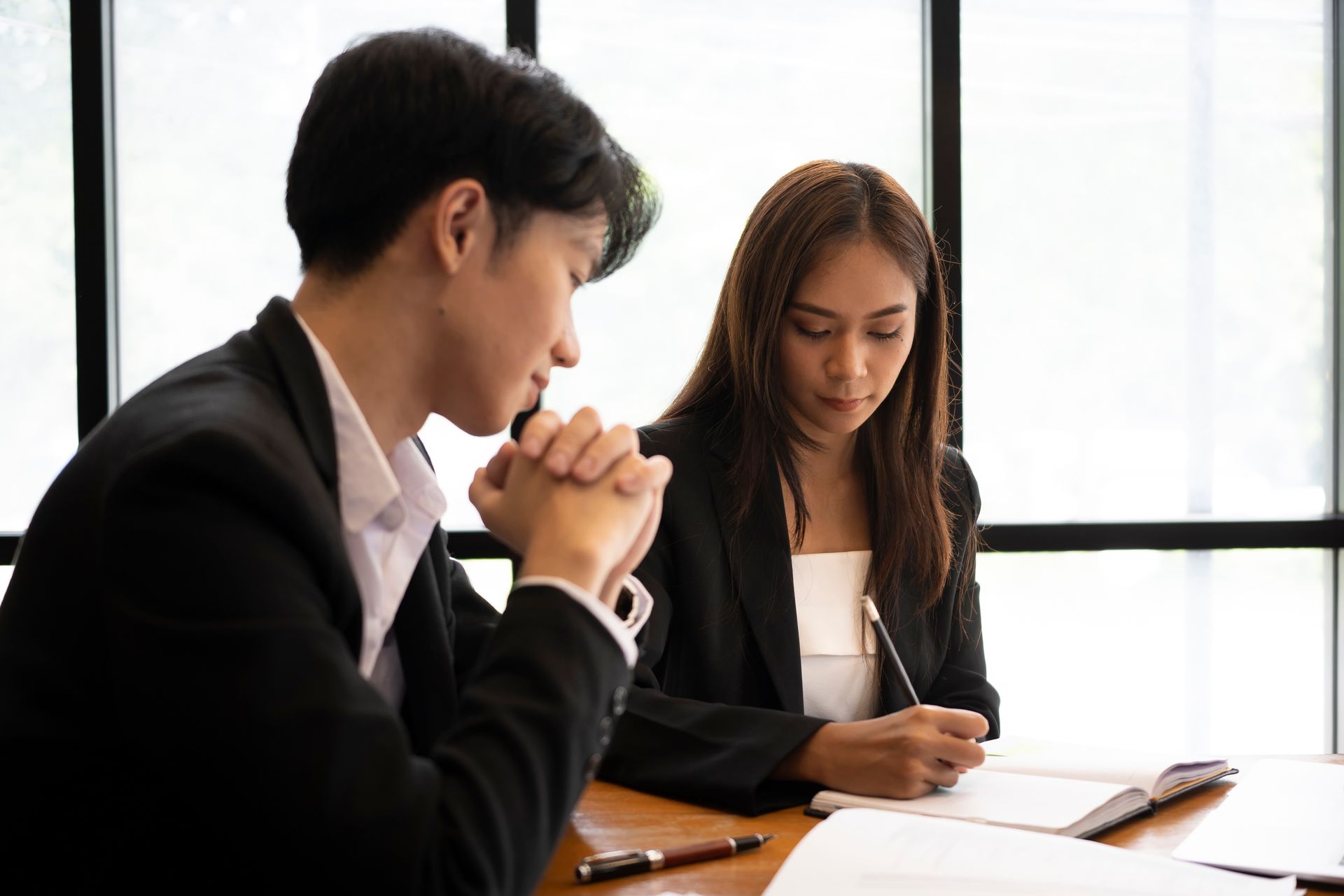 Two business professionals in dark suits seated at a table, one taking notes while the other listens with clasped hands.