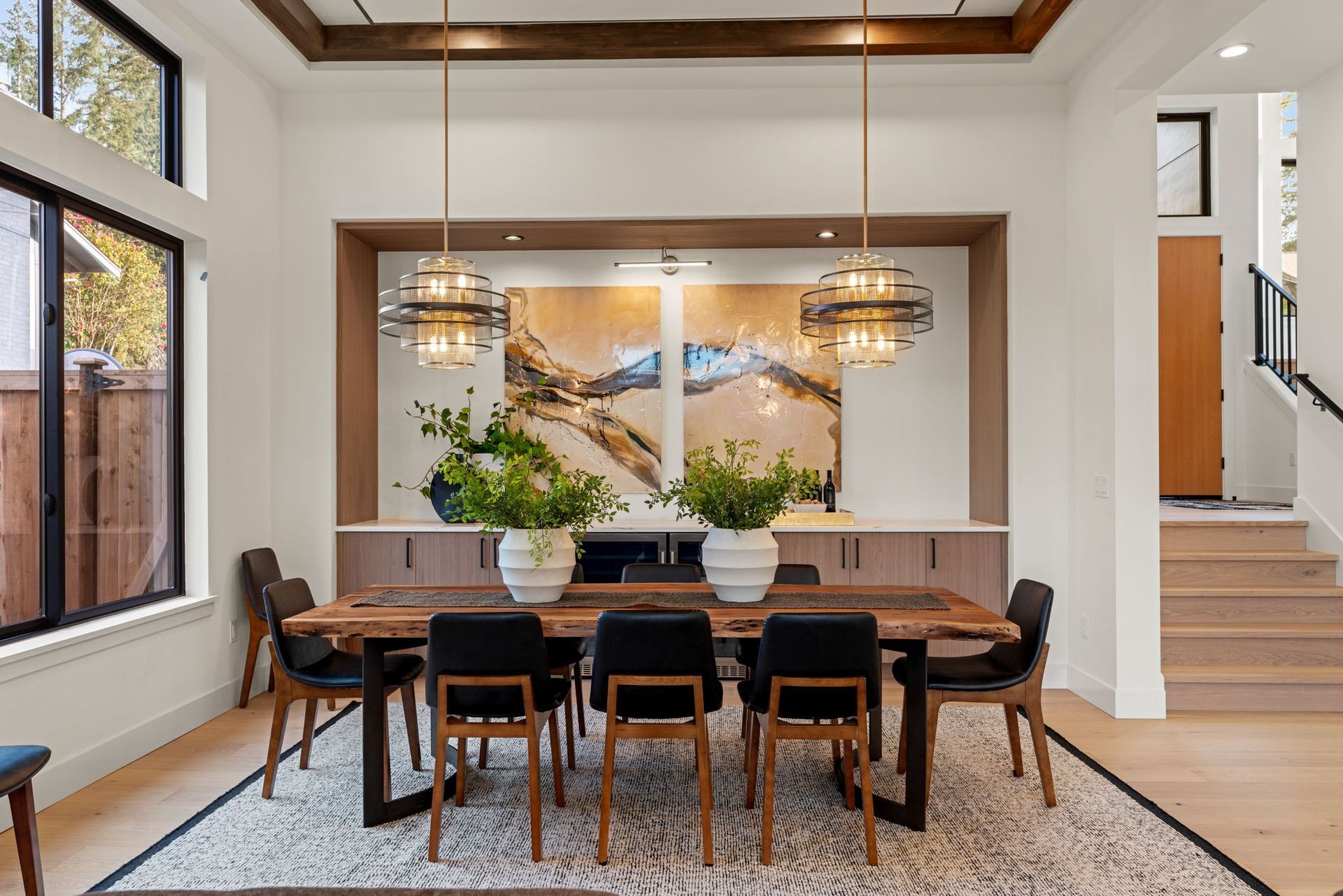 Dining room: wooden table, black chairs, artwork, two chandeliers, and a light-colored rug on a wood floor.