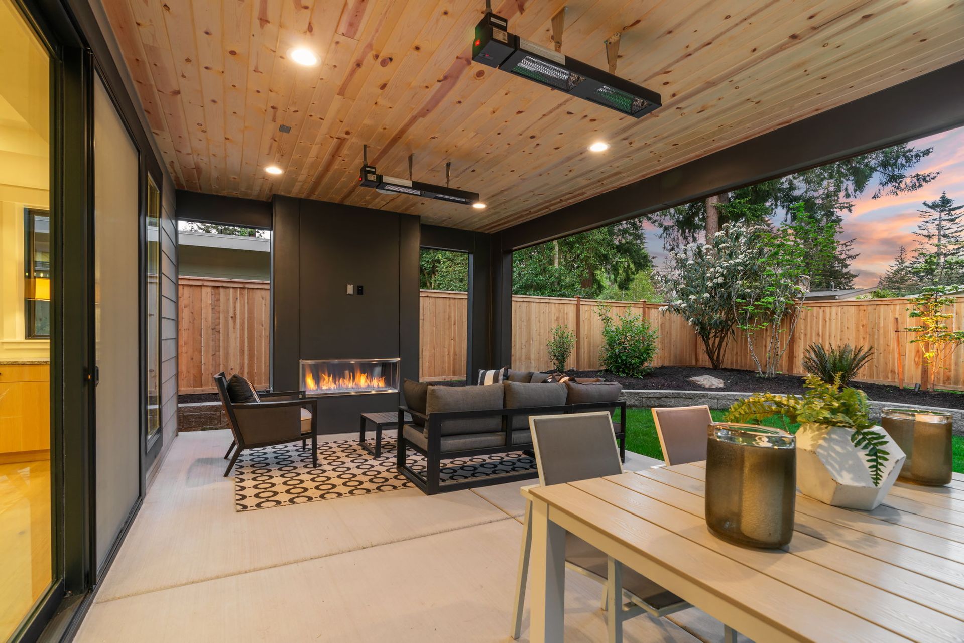 Covered patio with wicker seating, fireplace, wooden ceiling, and dining table beside greenery.