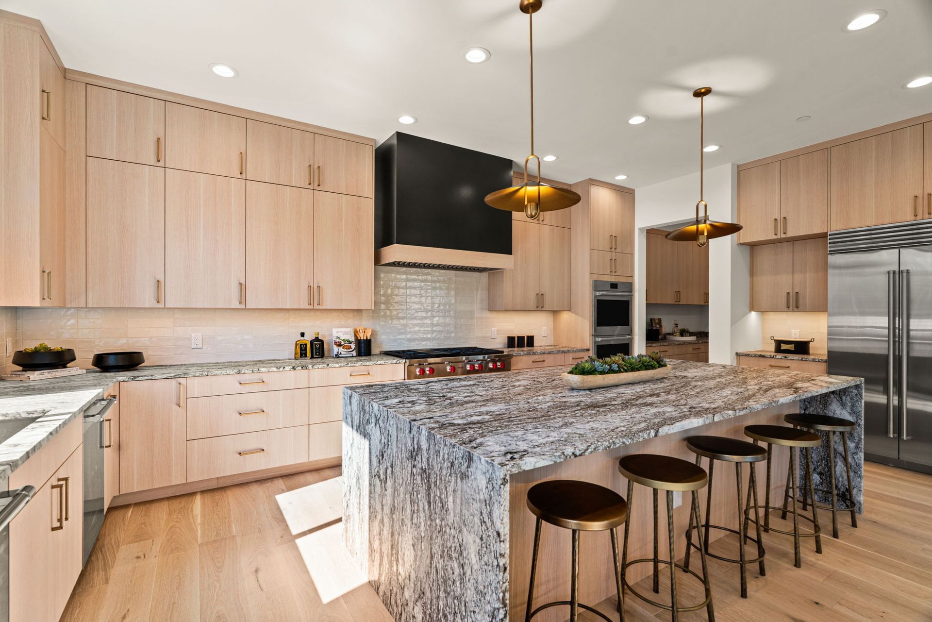 Modern kitchen with light wood cabinets, granite island, and four bar stools under pendant lights