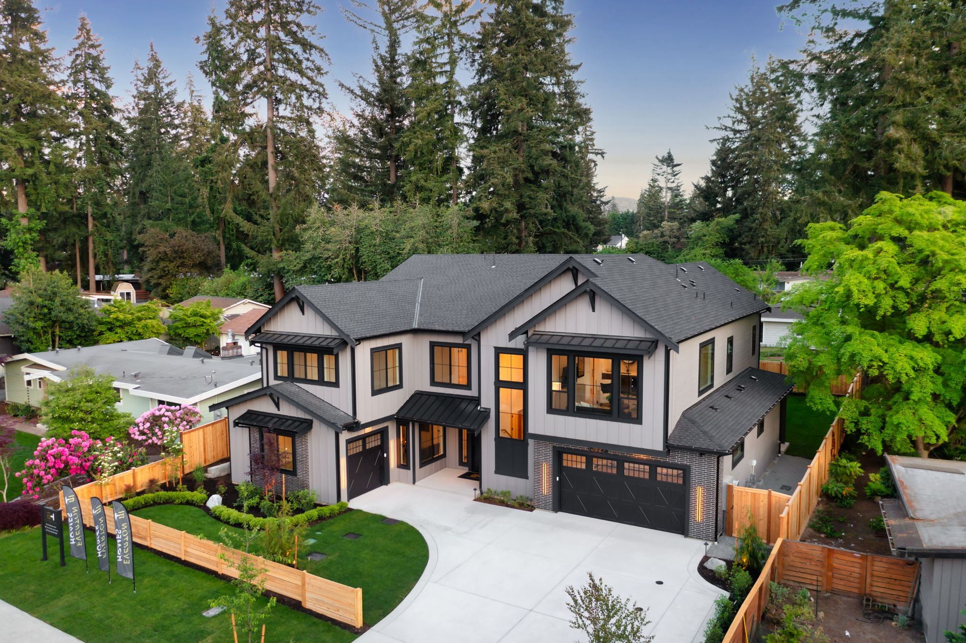 Large modern gray house with a driveway, lit windows, and tall trees in a landscaped yard at dusk