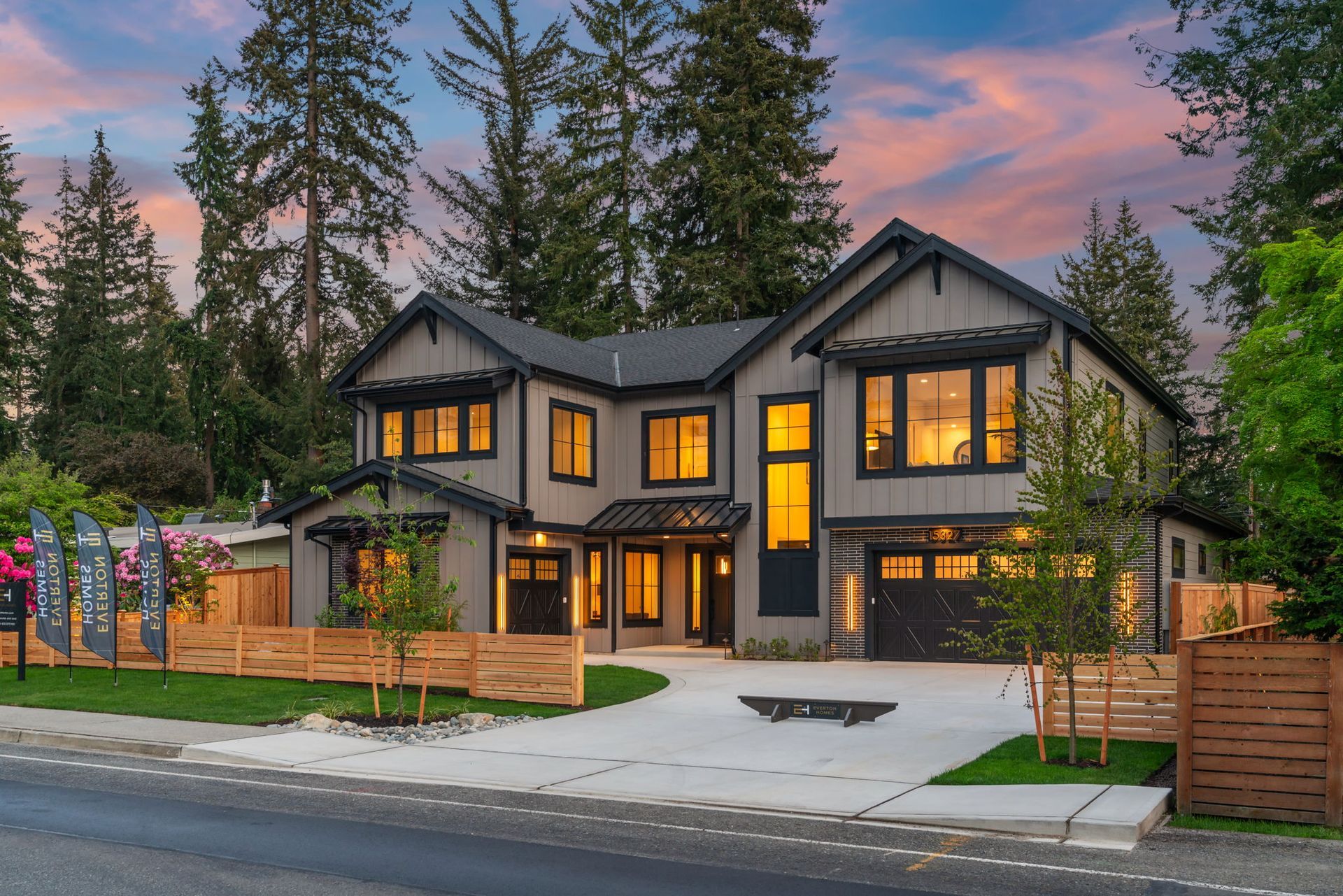 Modern two-story house with lit windows, large trees, and a pink sunset sky
