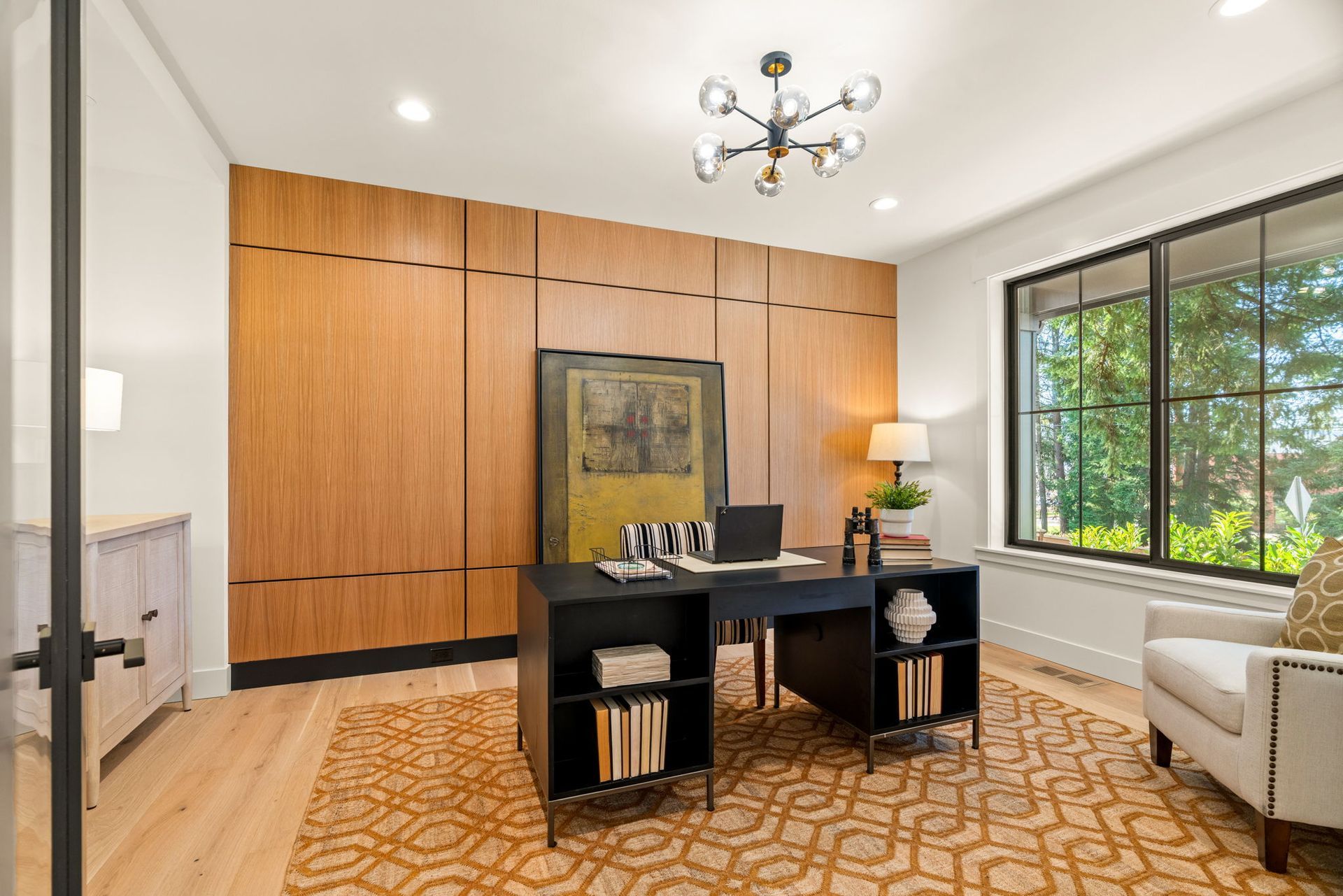 Home office with wood paneling, black desk, window, and patterned rug.