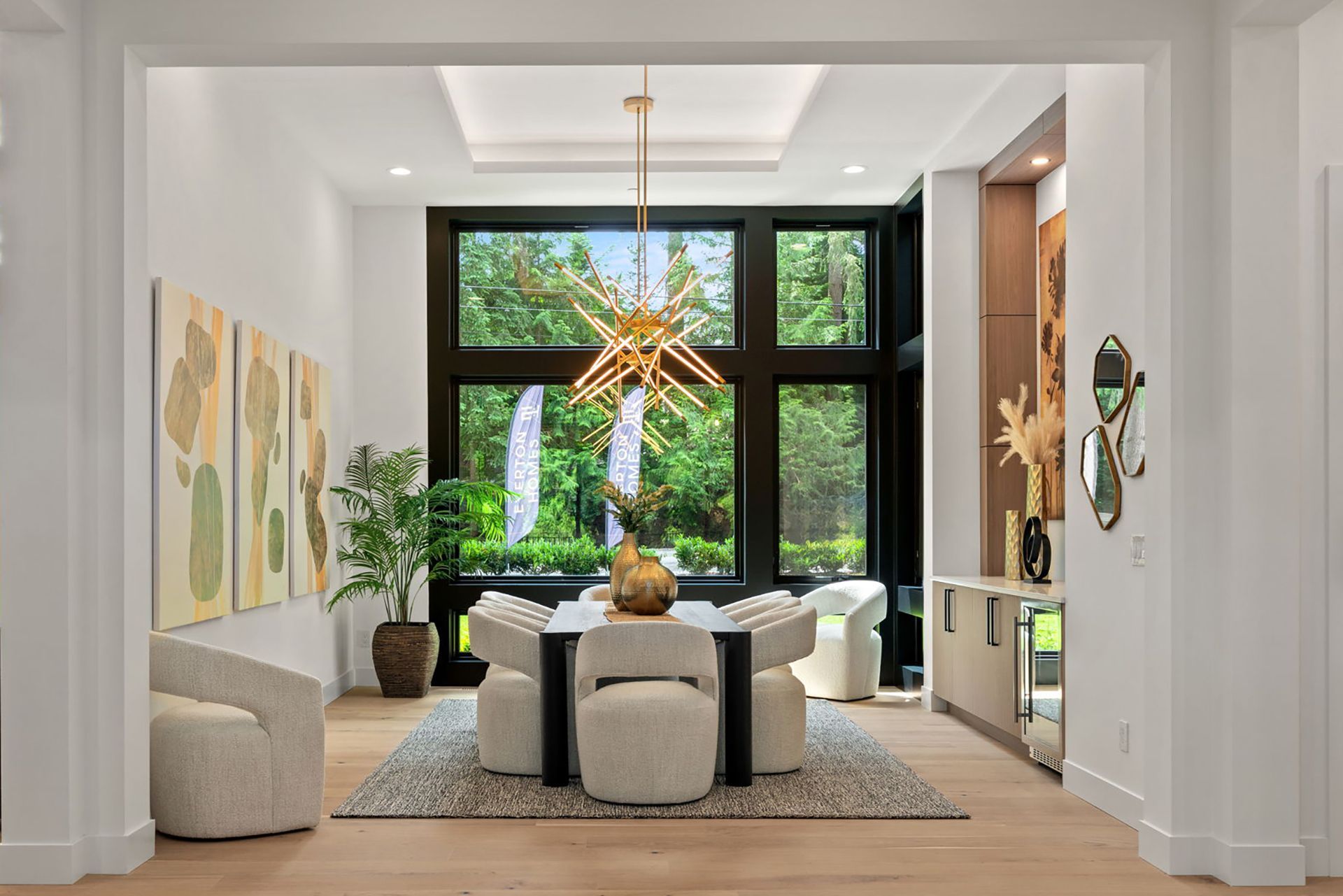 Elegant dining room with large window, round table, and gold chandelier. Artwork and greenery add ambiance.