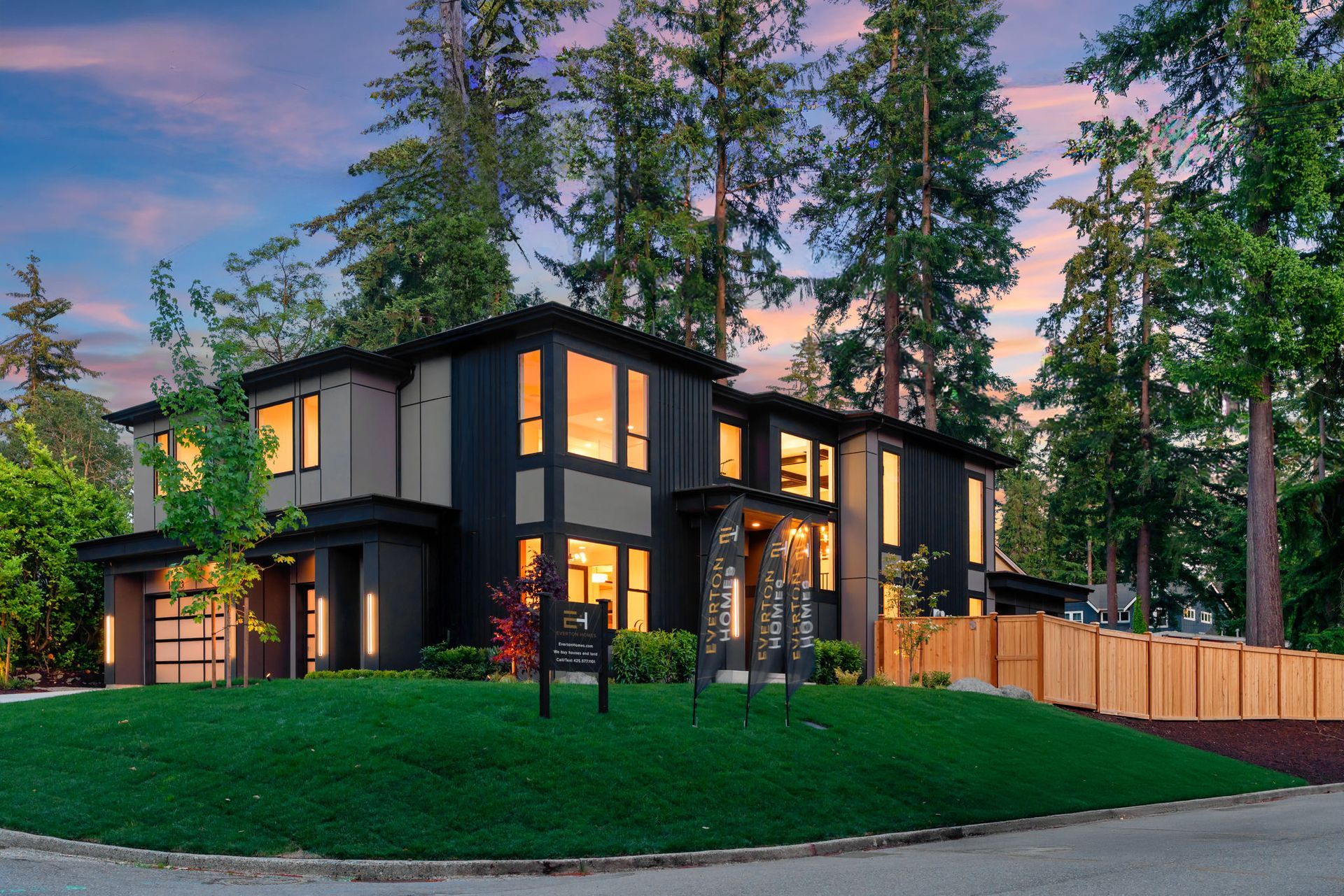 Modern two-story house with lit windows, tall trees, and a manicured lawn at dusk