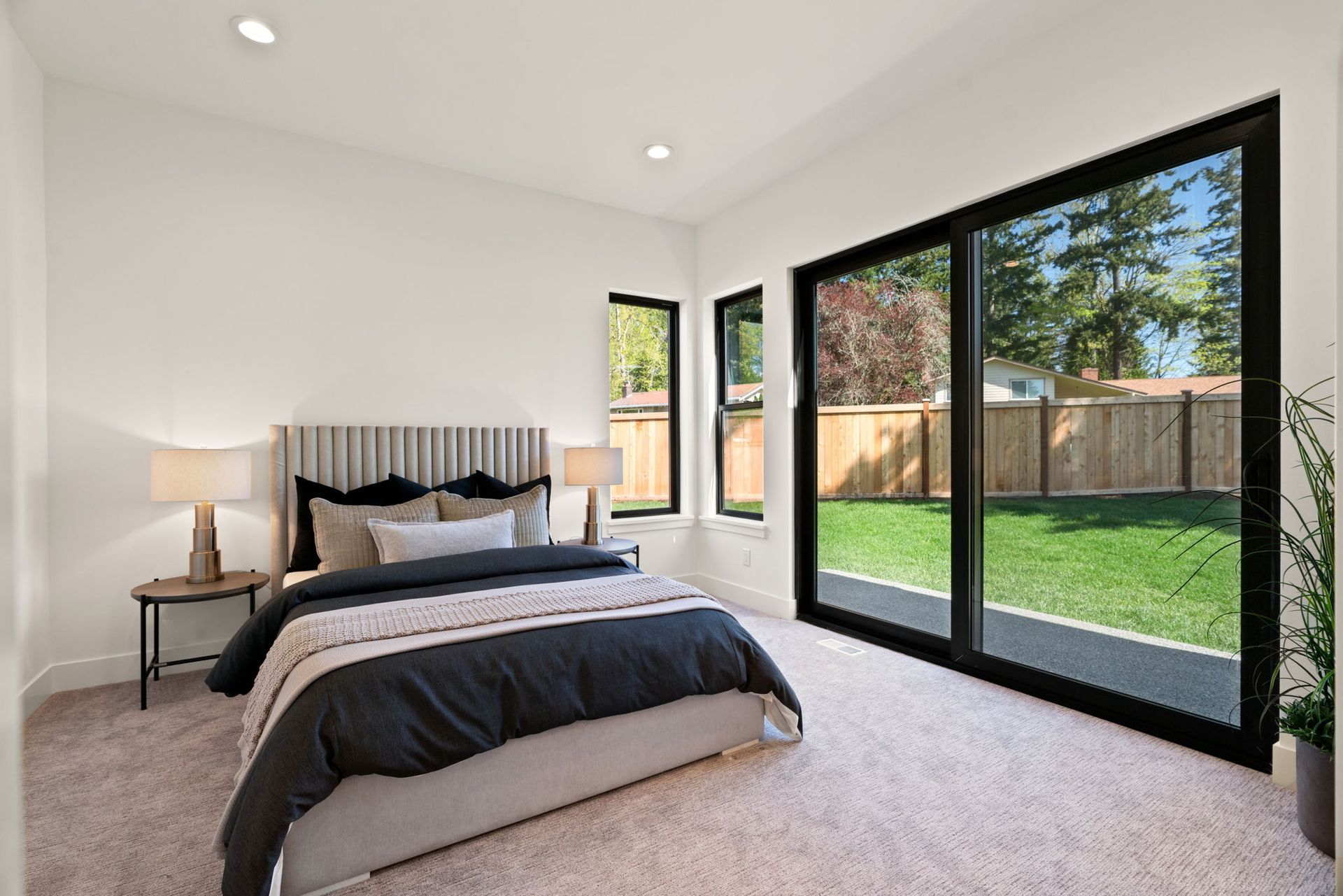 Bright bedroom with bed, nightstand, and large windows overlooking a green backyard.