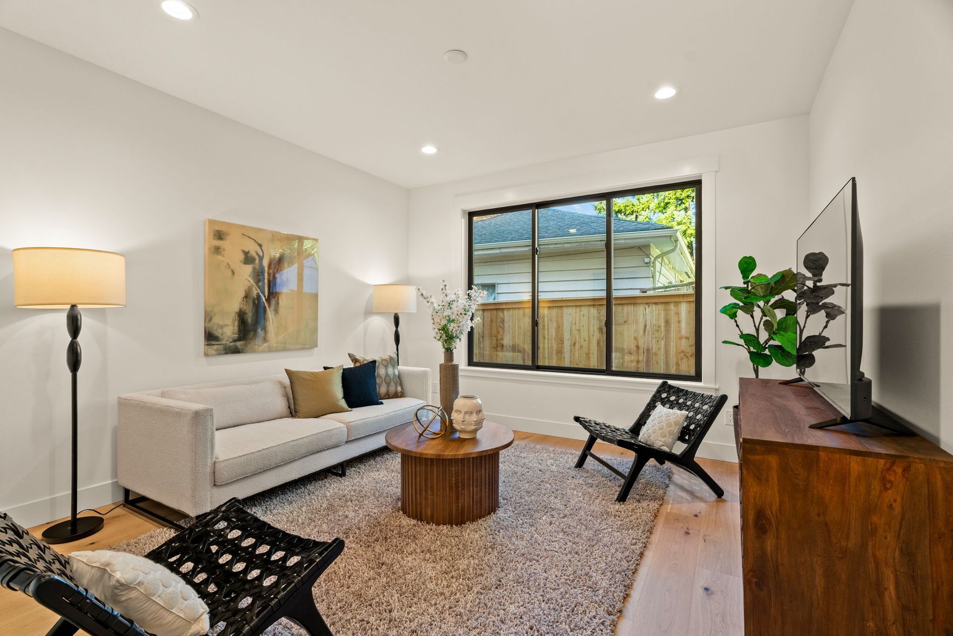 Living room with a beige sofa, two black chairs, round wooden table, and a large window.