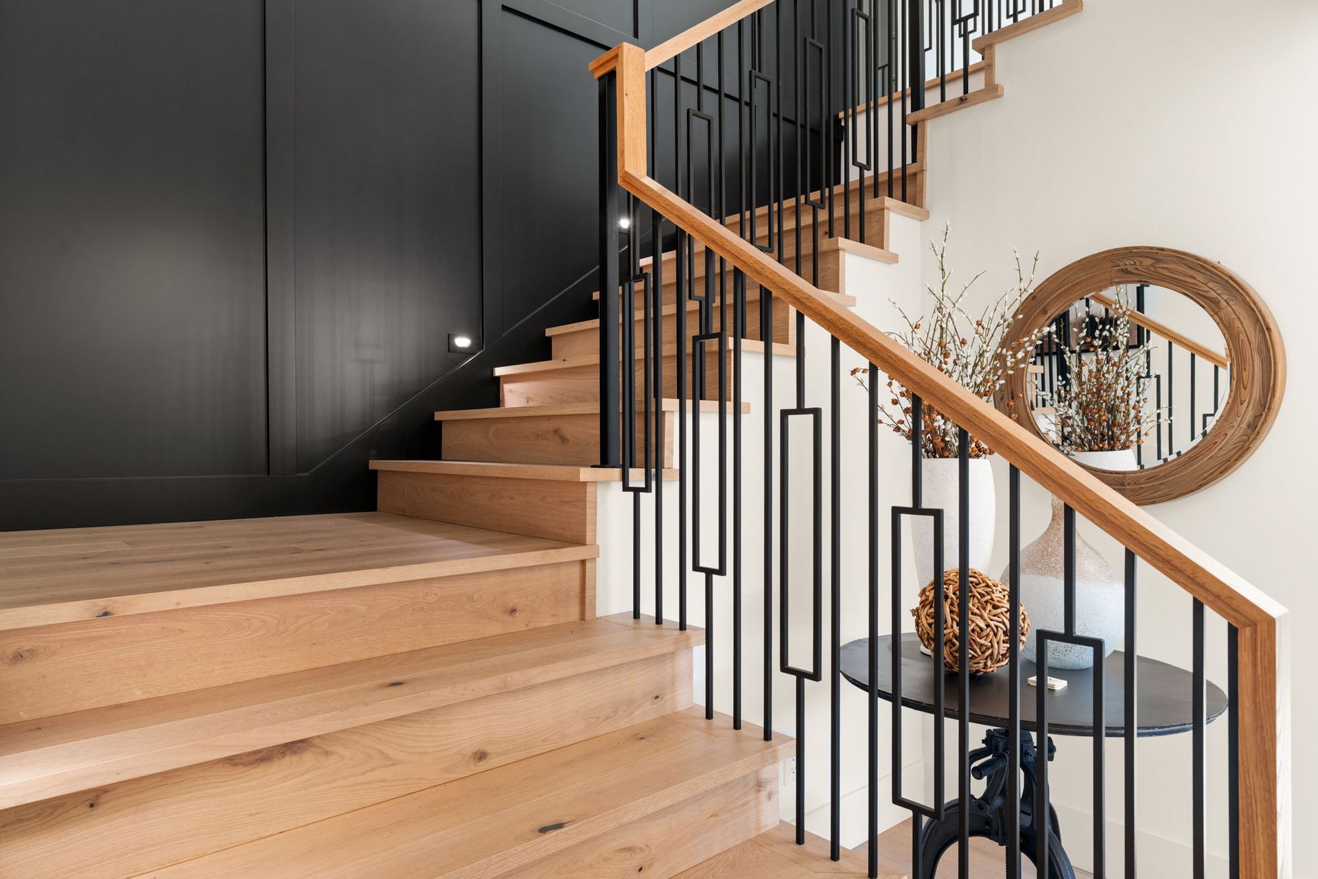 Wooden staircase with black and light wood details, round mirror, table, and decorative vase.