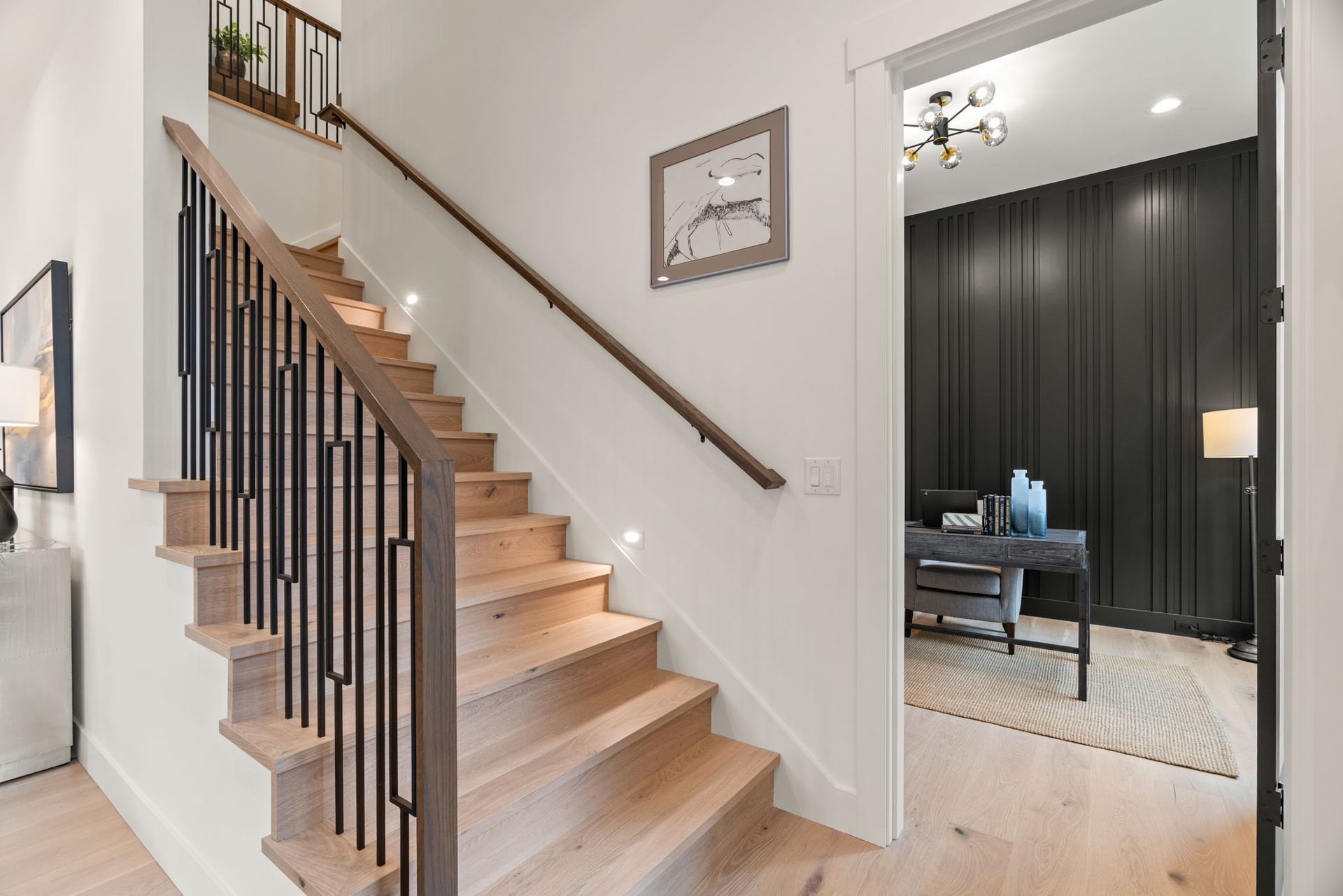 Wooden staircase with black metal railings, leading up to a landing. White walls and an office in the background.