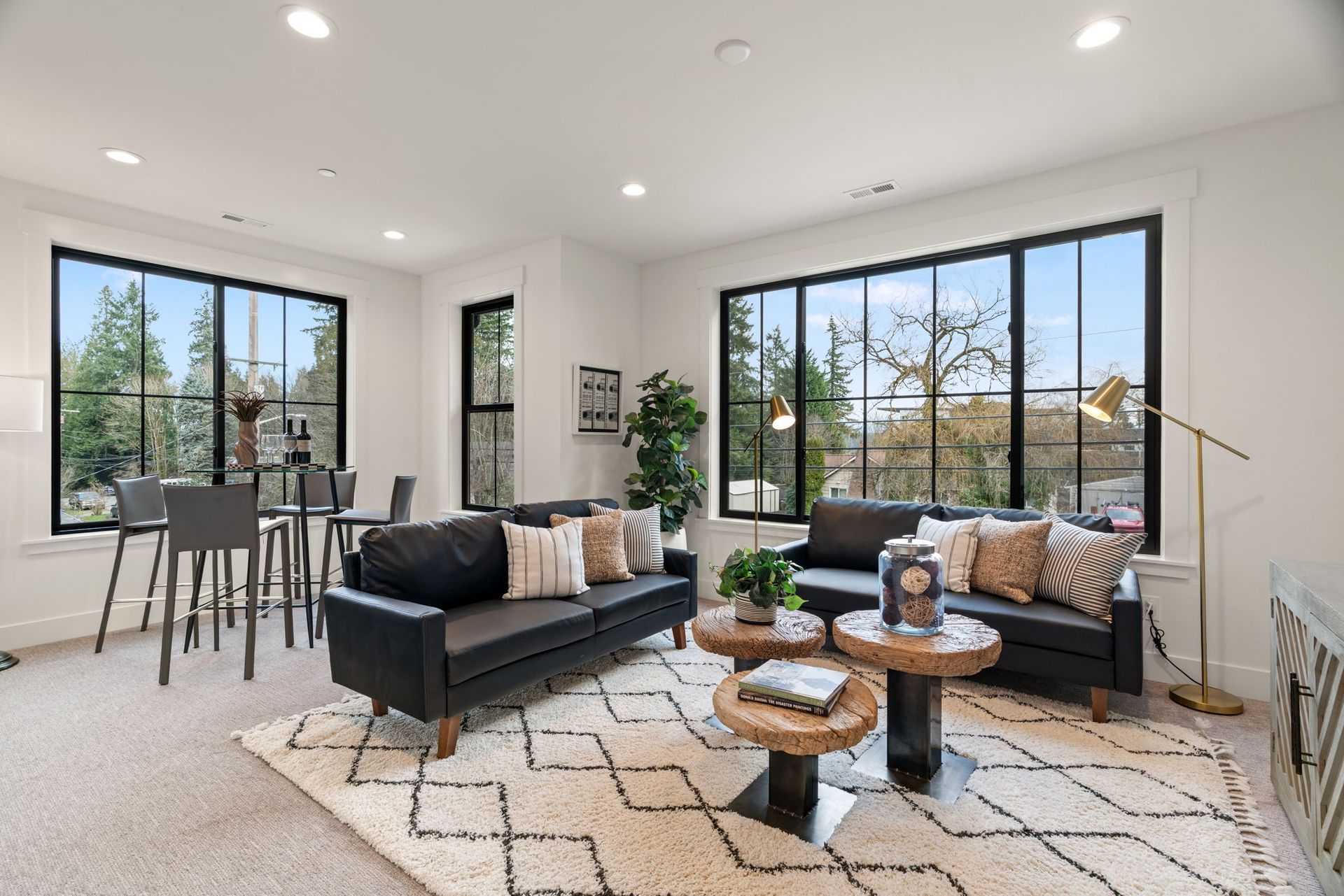 Living room with black-framed windows, dark sofas, coffee tables, area rug, and bar stools. Bright, airy space with outdoor view.