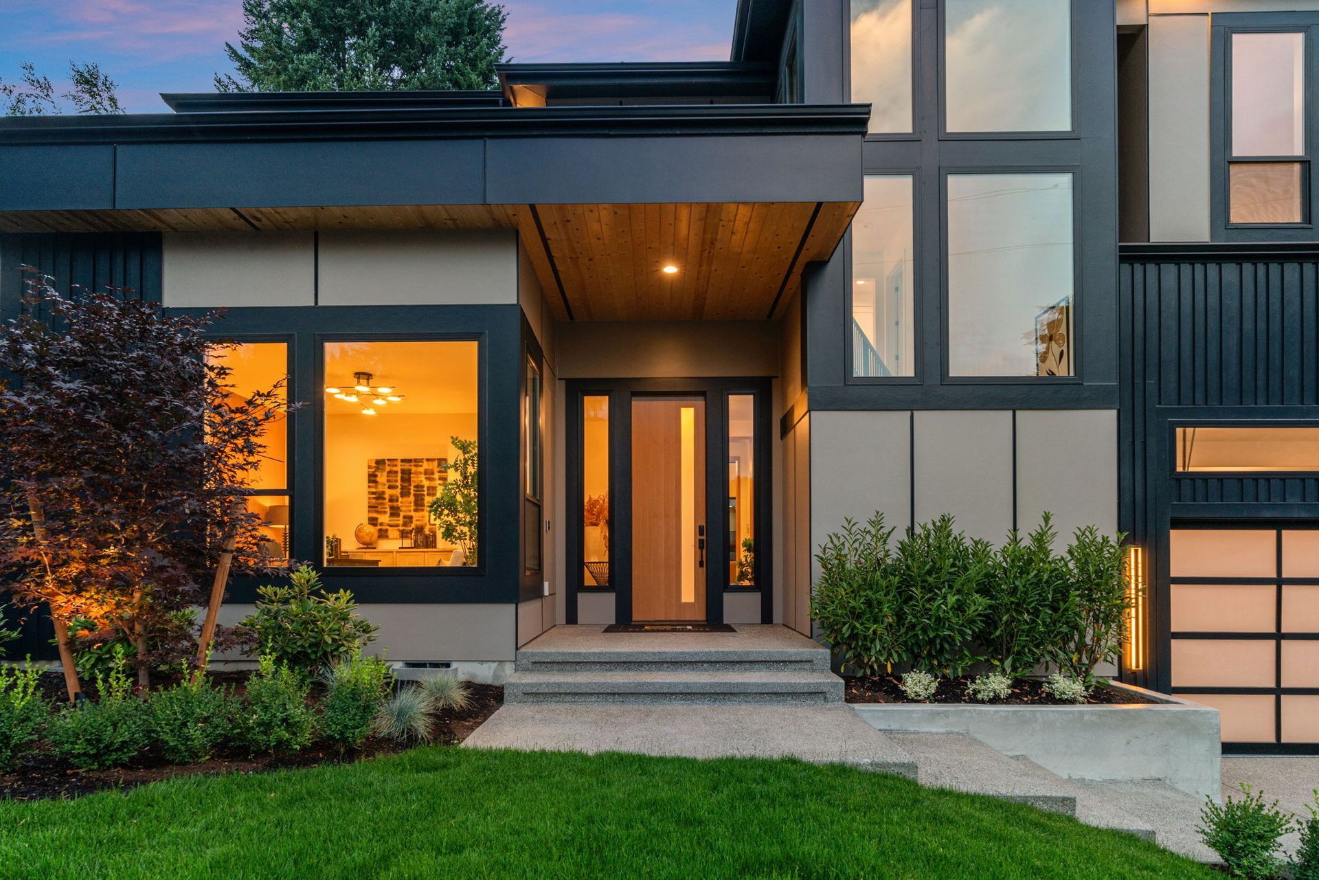 Modern house exterior at dusk with lit windows, front entry steps, and landscaped lawn
