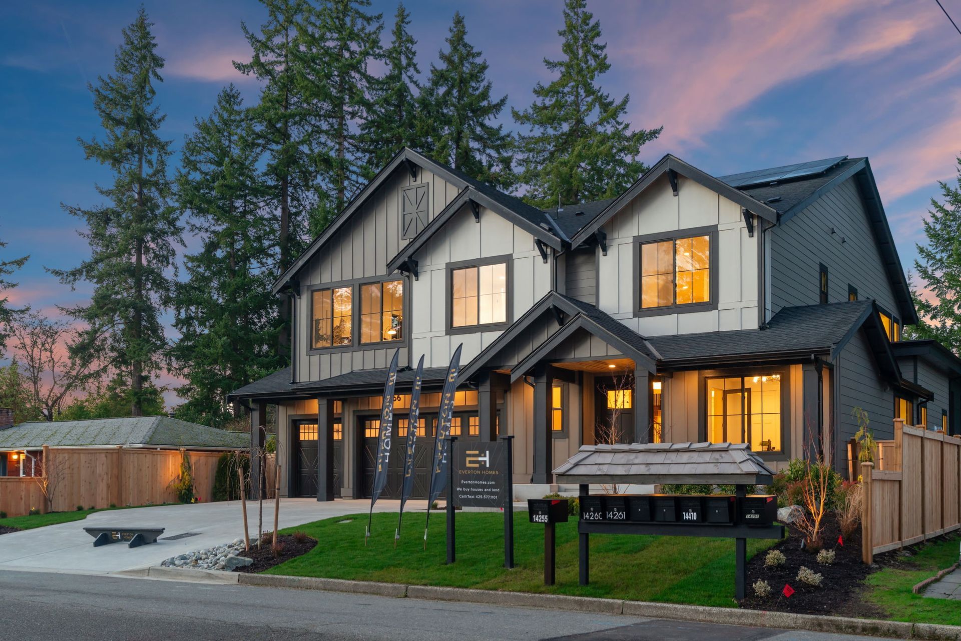 Modern two-story house with dark gray trim and light siding, lit windows, and a landscaped yard at dusk.