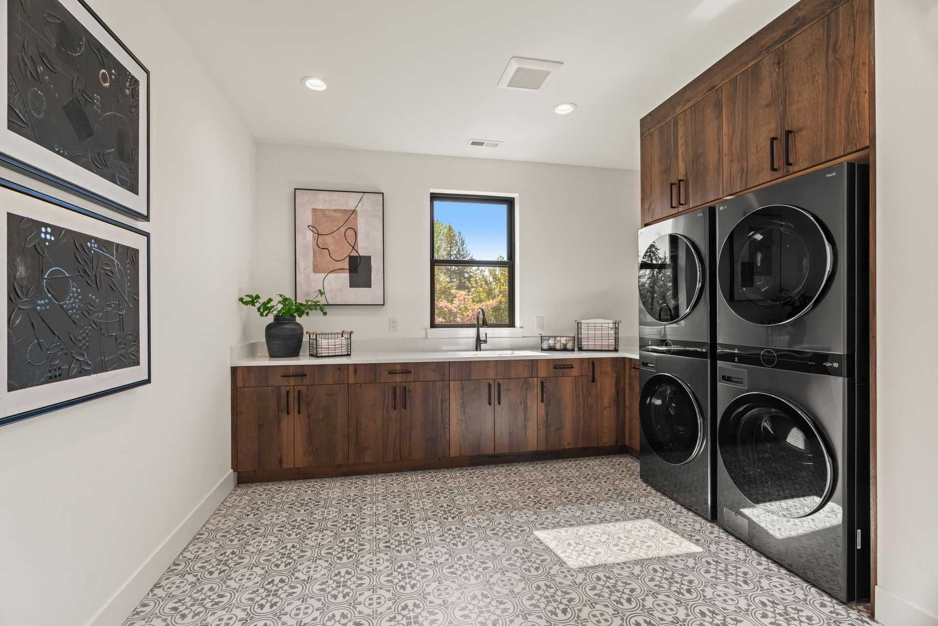 Laundry room with wood cabinets, stacked black washer-dryer units, and a window above a sink.