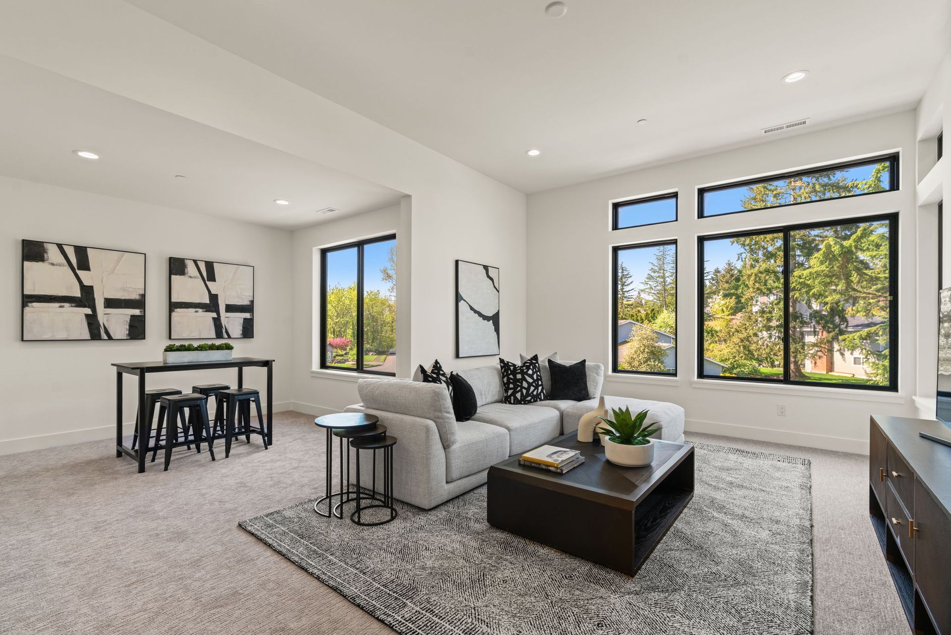 Modern living room with gray sofa, black coffee table, dining nook, and large windows overlooking trees