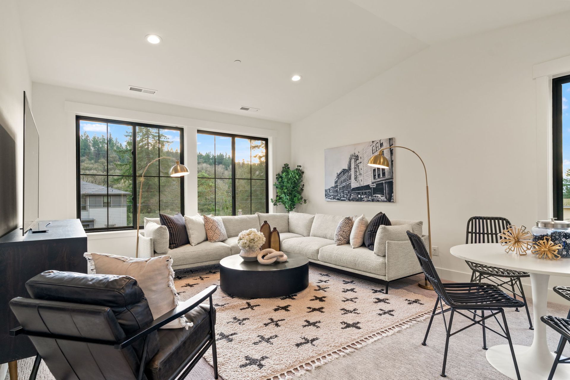Living room with white walls, sectional sofa, and round coffee table on a patterned rug near large windows.