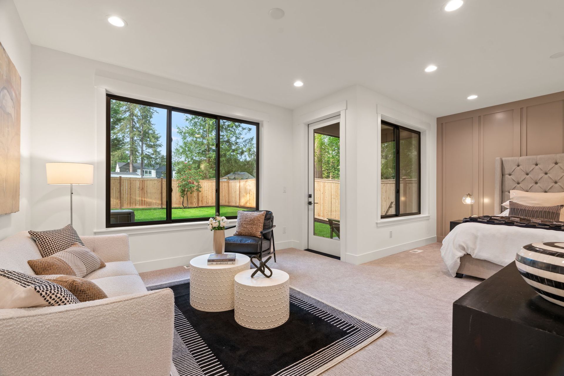 Bright modern bedroom with large windows, beige bed, sitting area, and patterned rug