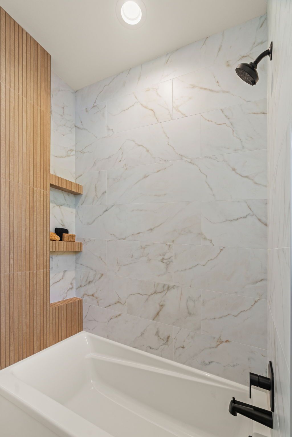Small modern bathroom with white marble-look walls, a bathtub, wood slat accent, and black shower fixtures