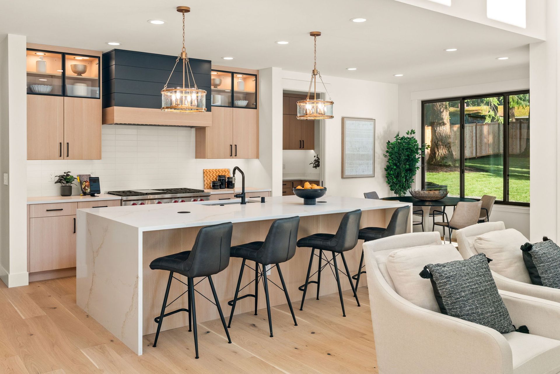Modern kitchen with island, seating, and natural light. Light wood cabinets, white countertops, and black stools.