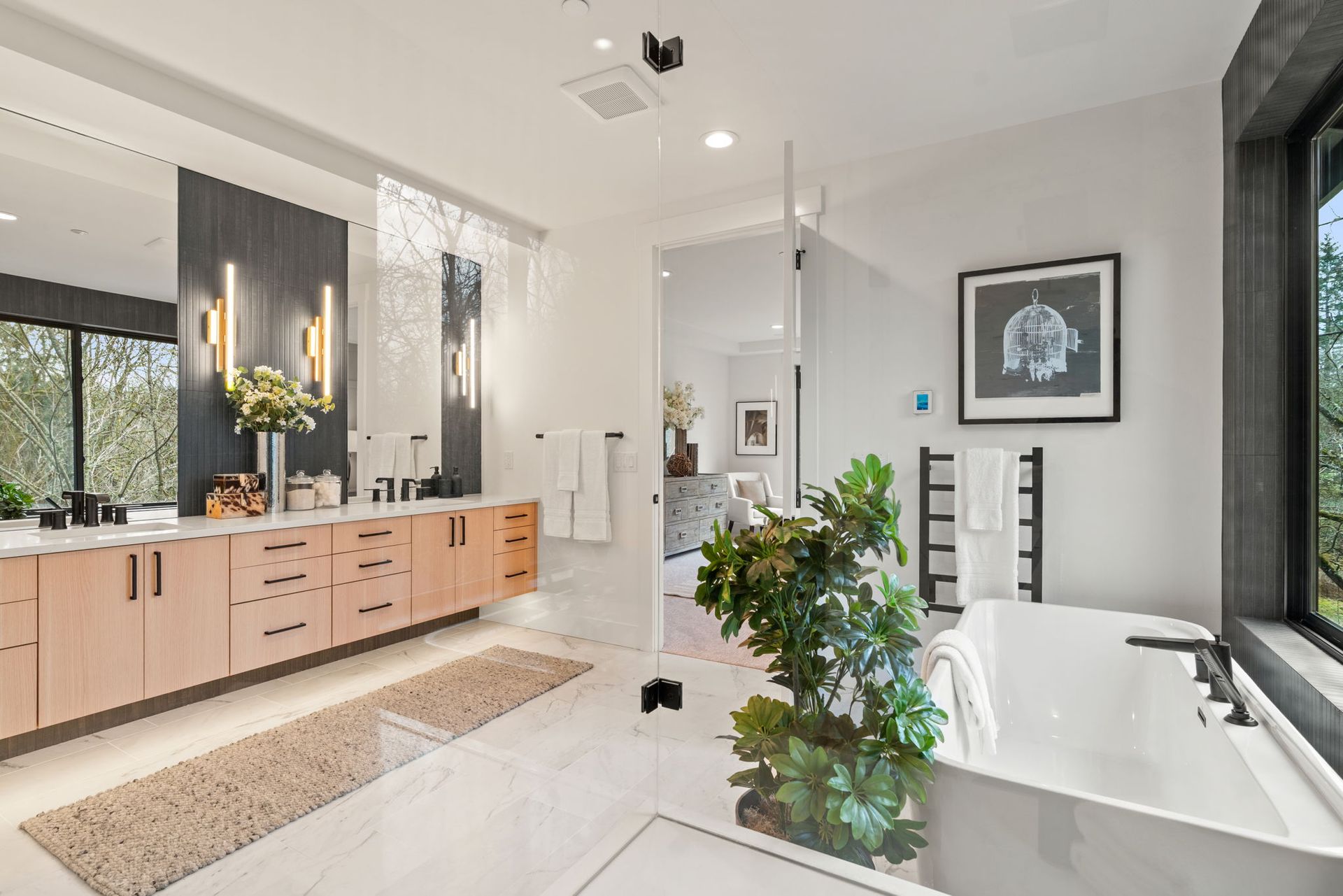 Modern bathroom with light wood cabinets, white walls, freestanding tub, and a large window with a view.