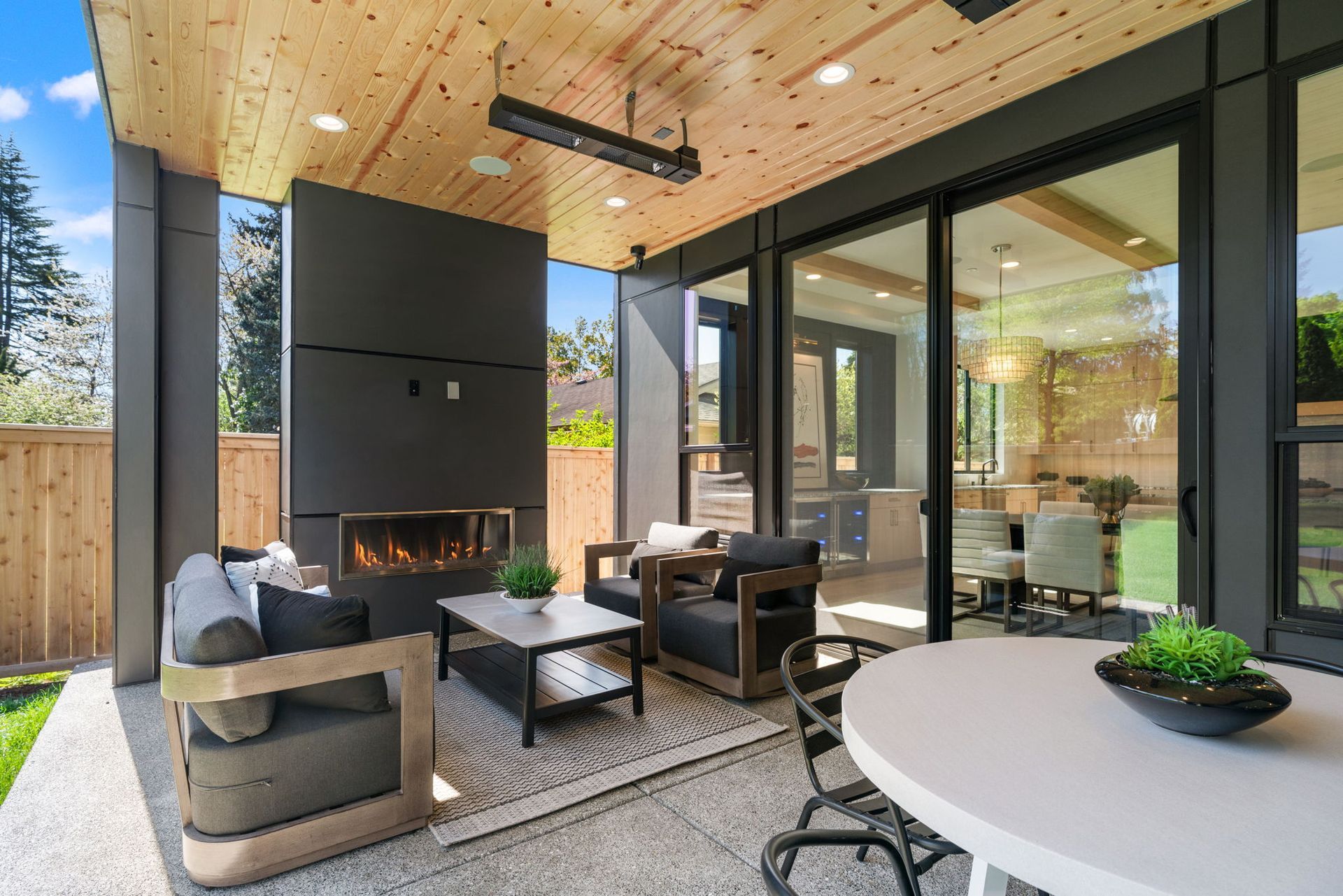 Modern covered patio with fireplace, lounge chairs, and dining table beside glass walls and wood ceiling