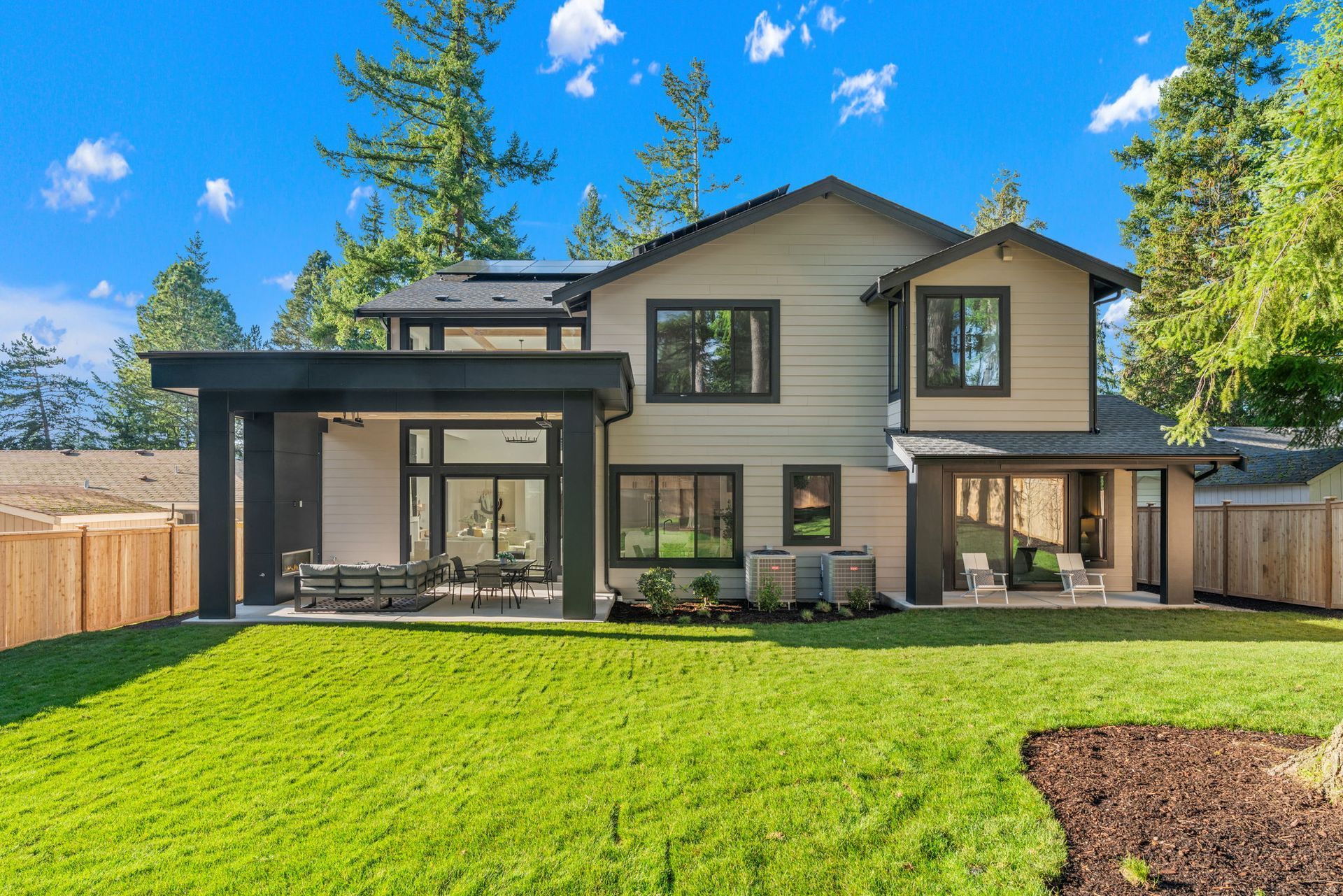 Rear exterior of a modern house with a green lawn, patio, and wooden fence on a sunny day.