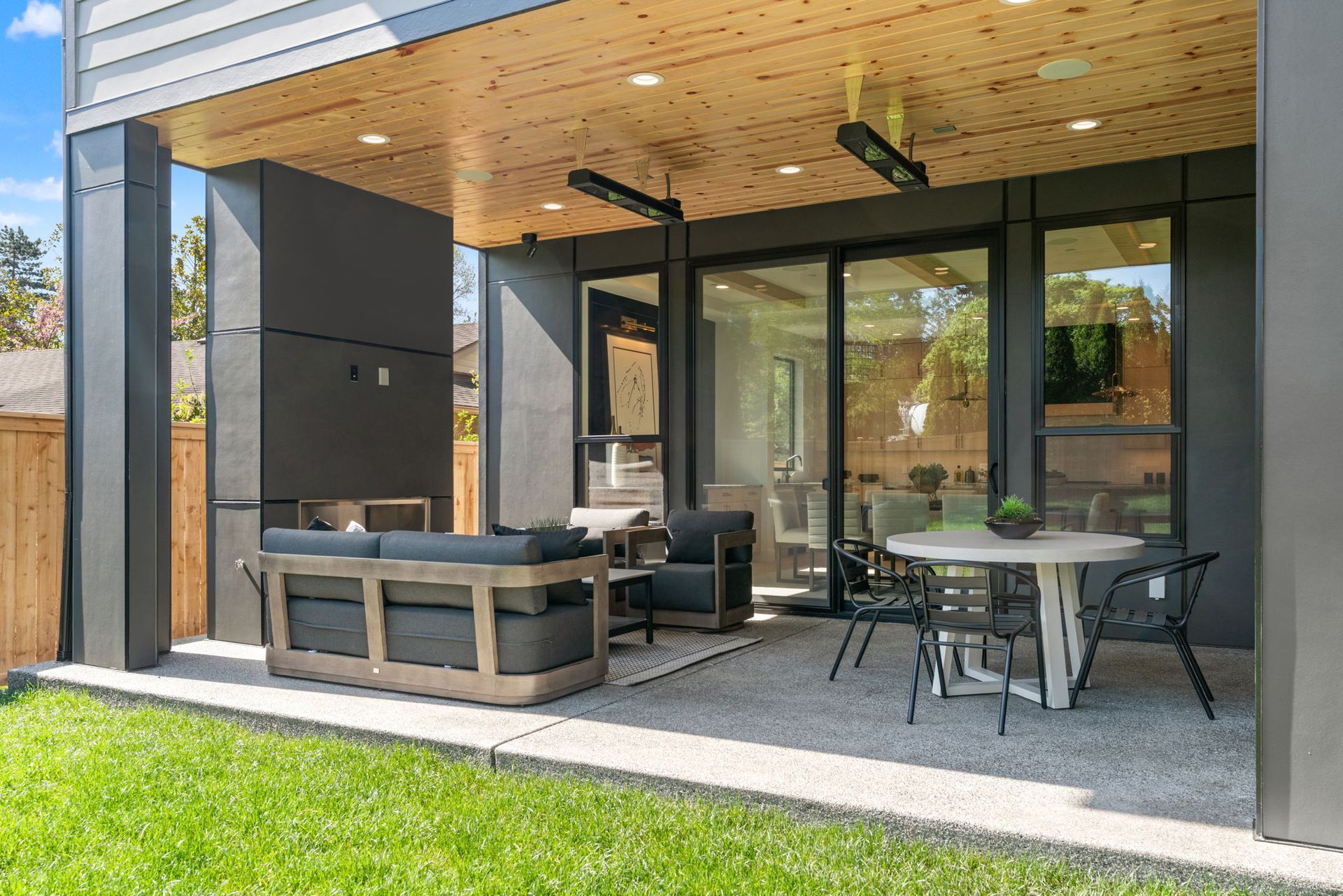 Modern patio with gray sectional seating, round table, and glass doors under a wooden overhang.