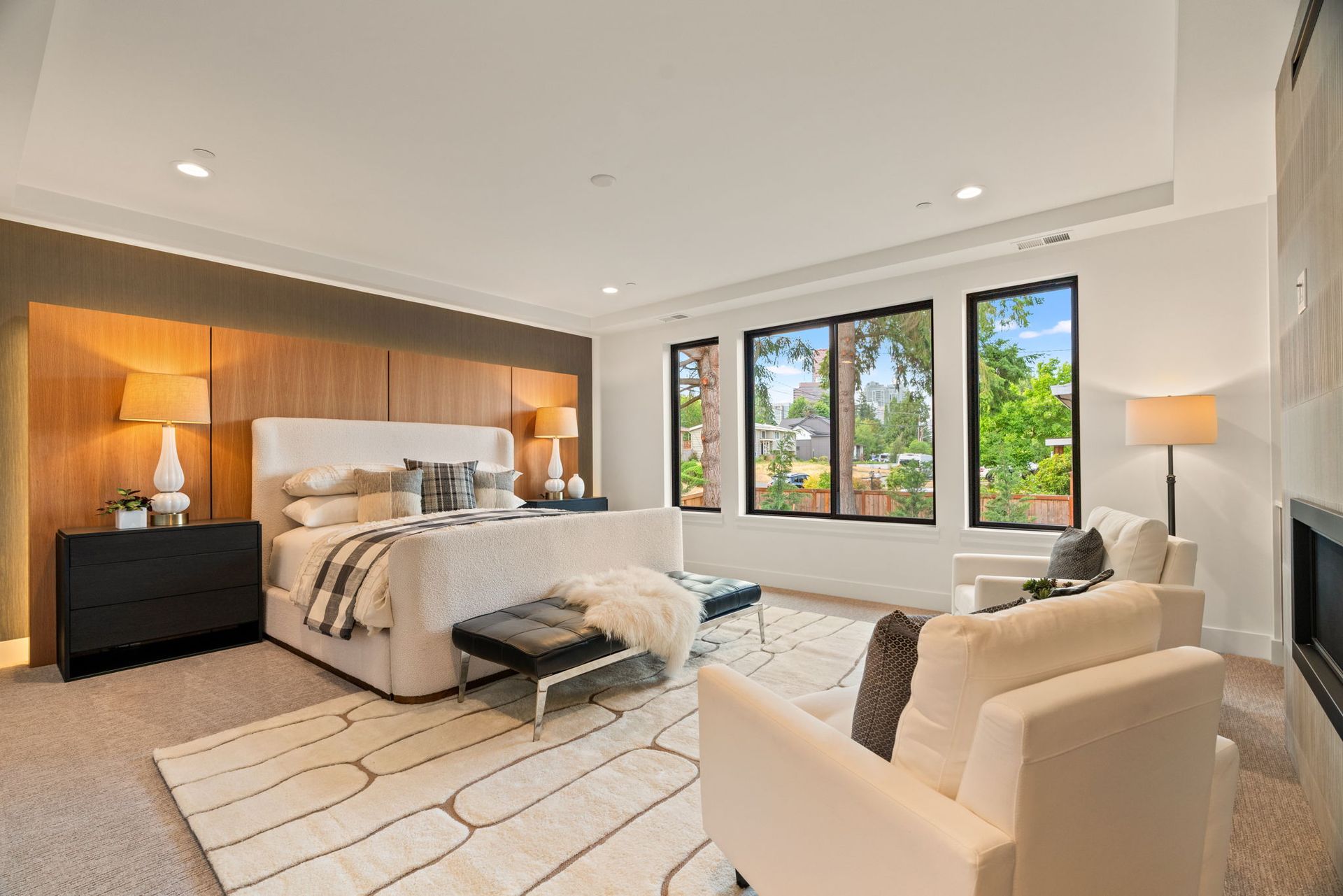Modern, primary bedroom with a large bed, wood accent wall, lounge chair, and bright windows overlooking greenery