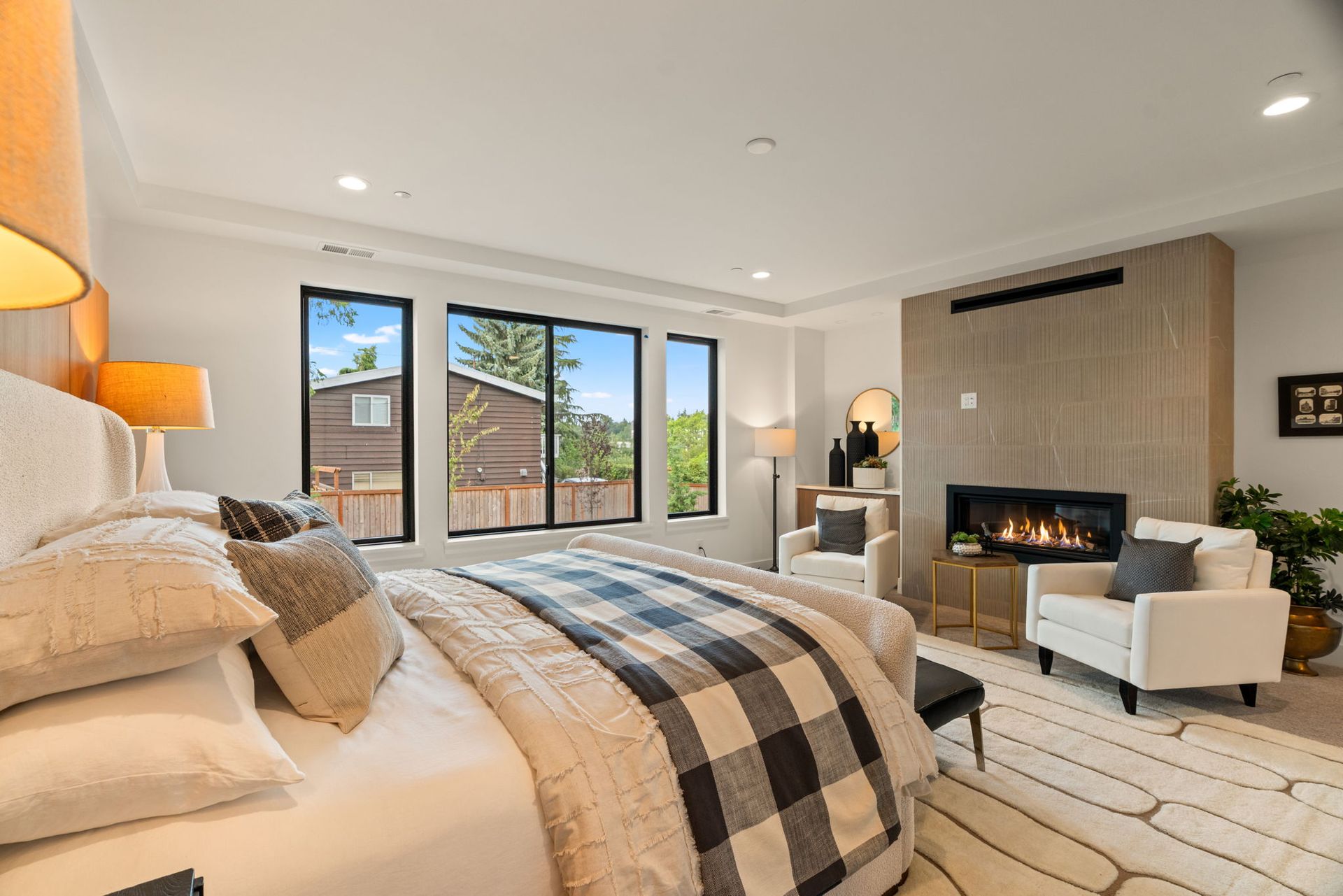 Modern bedroom with a fireplace, large windows, beige decor, and a plaid bedspread.
