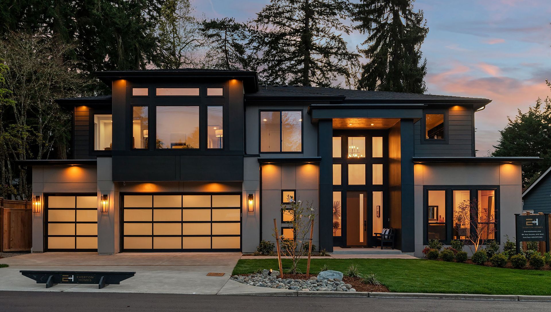 Modern two-story house with gray exterior, large windows, and garage doors, lawn, and cloudy sunset sky.