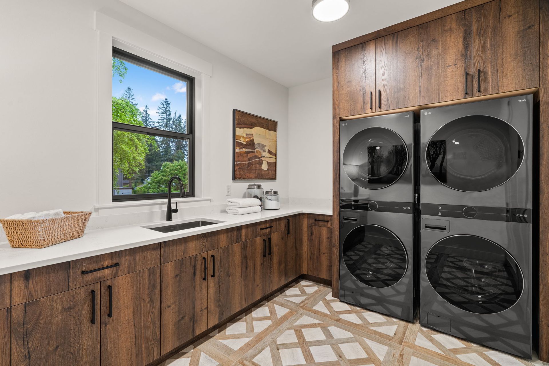 Modern laundry room with wood cabinets, white counters, sink, and stacked washer-dryer under a window