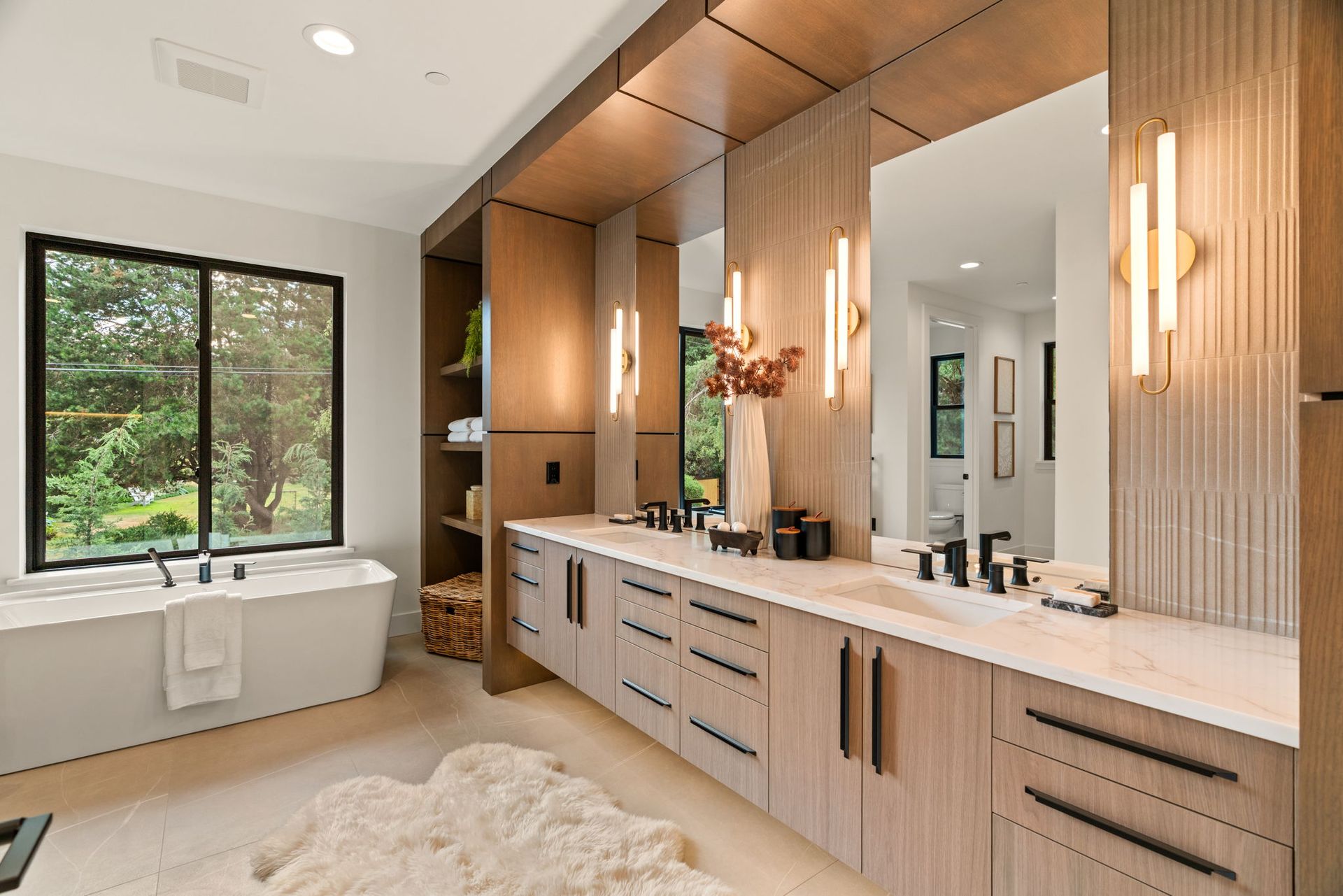 Modern bathroom with double vanity, large mirror, wood cabinetry, and a window beside a soaking tub