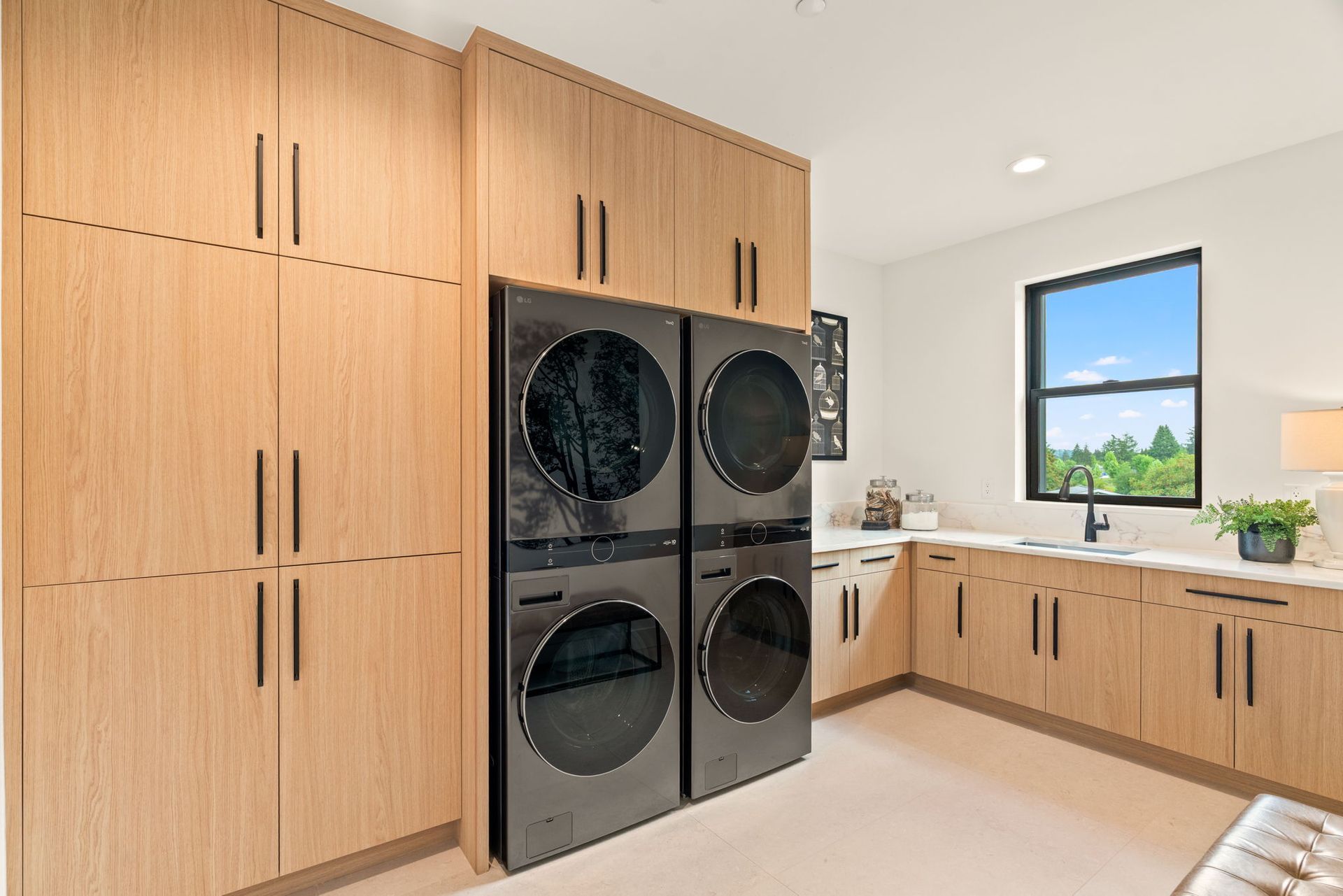 Laundry room with dual, stacked, black washer and dryer, light wood cabinets, and a countertop by a window.