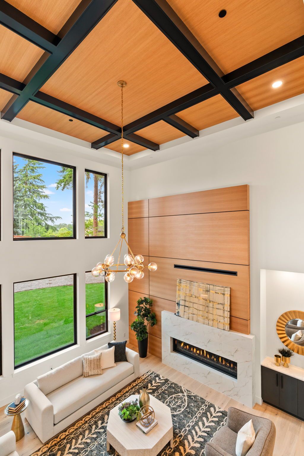 Bright, modern, two-story great room with vaulted wood ceiling, white walls, fireplace, sofa, and large windows overlooking greenery